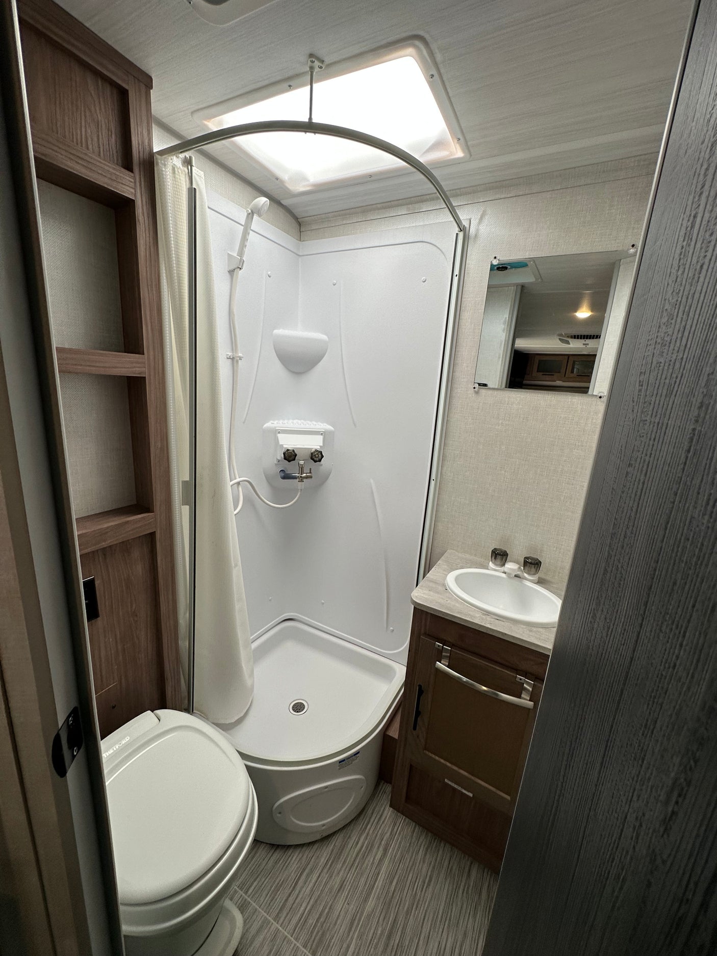 Clean and compact RV rental bathroom: shower, toilet, sink vanity, mirror, and shelving. Brightly lit and functional design.