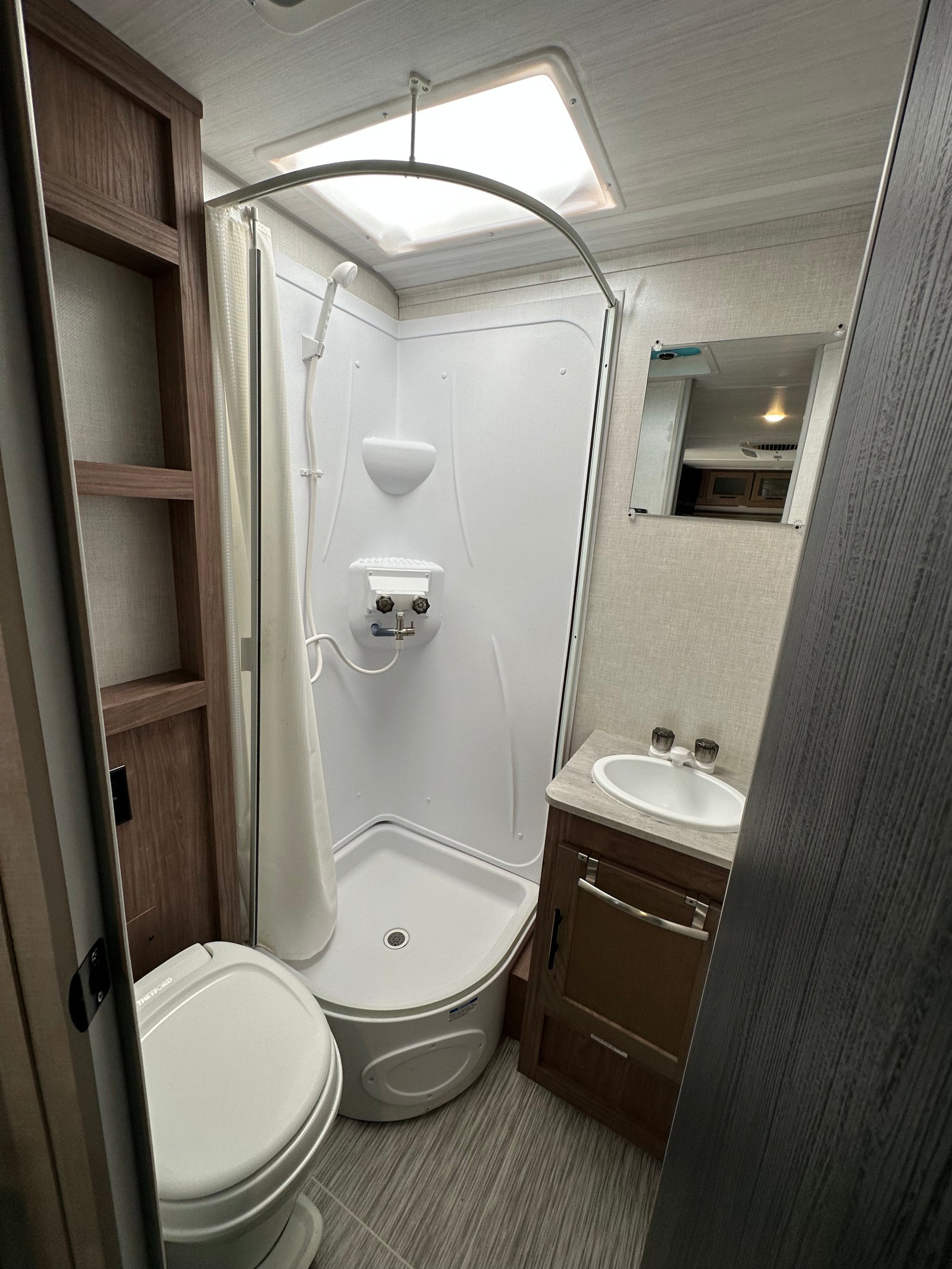 Clean and compact RV rental bathroom: shower, toilet, sink vanity, mirror, and shelving. Brightly lit and functional design.