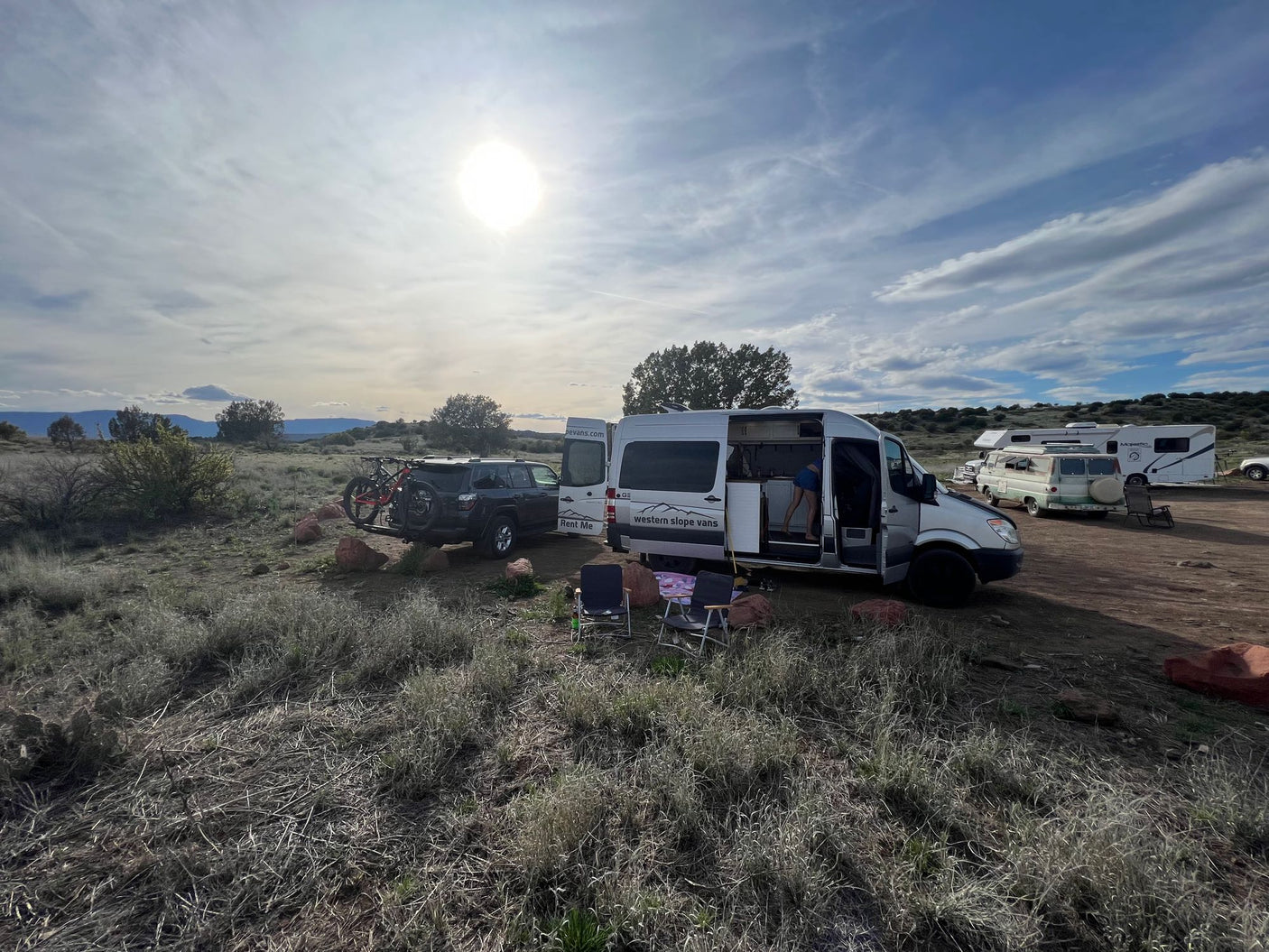 Enjoy adventure travel with an RV rental! White vans & SUV at a dry, open campsite with chairs, bikes, and mountains.