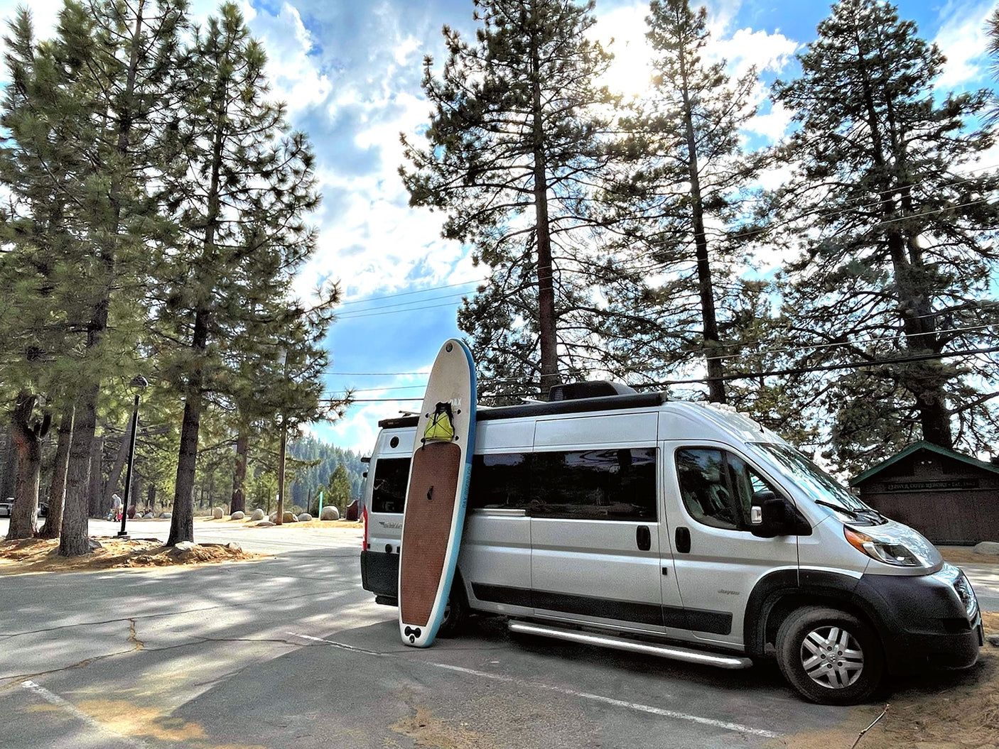 An rv rental camper van with a paddleboard is parked under tall pine trees, perfect for a sunny outdoor adventure.