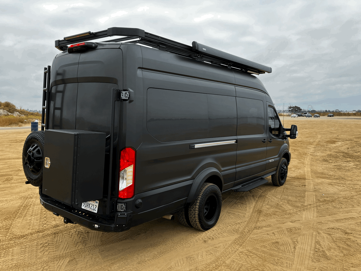 Black Ford Transit RV rental camper van on sand, with roof rack & awning.