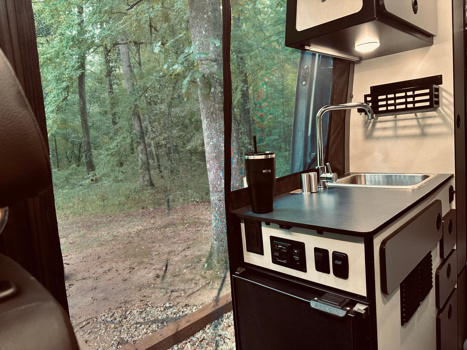Inside an RV rental: a kitchenette with sink, controls, and fridge, looking out to a serene forest scene through a screen. Perfect for camping!