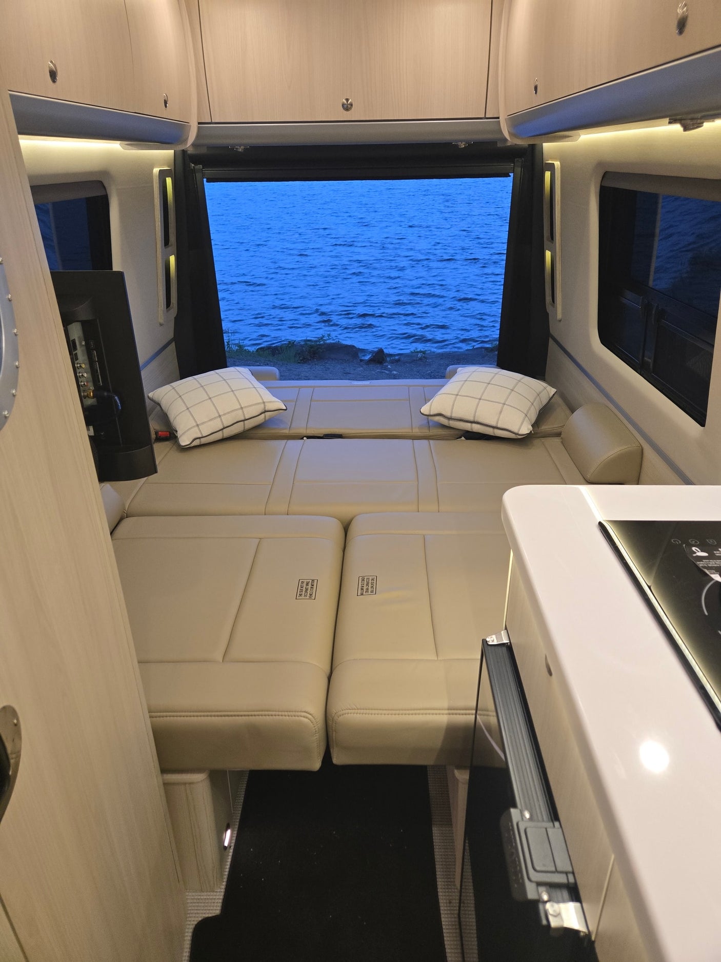 Interior of a modern rv rental shows a cream upholstered bed, compact kitchen counter, and open doors revealing a tranquil blue lake.