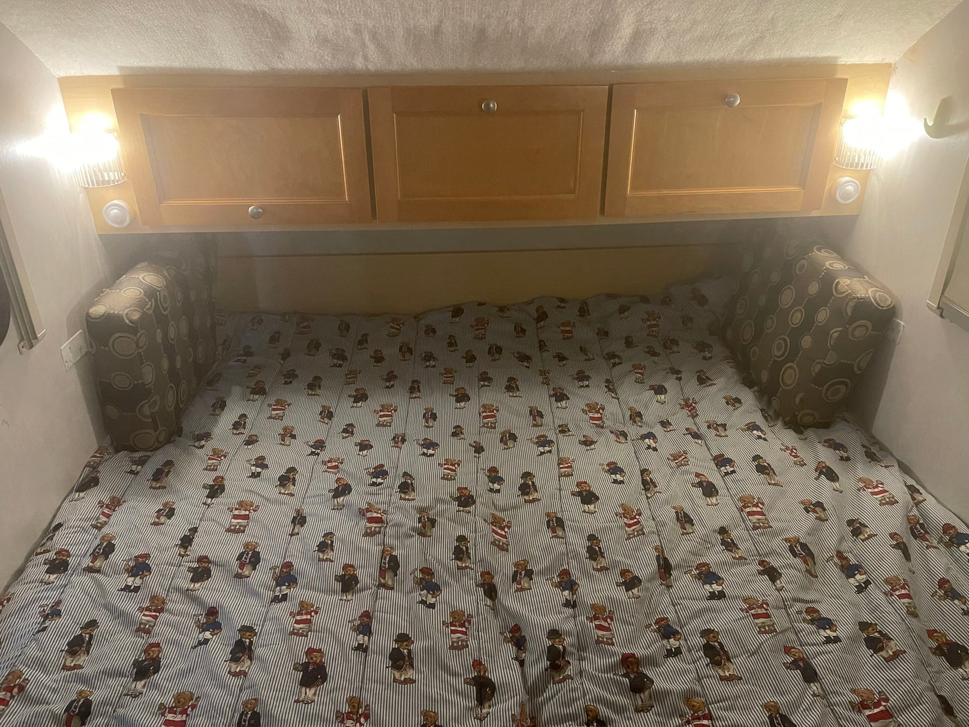 Inside an RV rental: a cozy bed with teddy bear striped comforter, overhead cabinets, and reading lights.