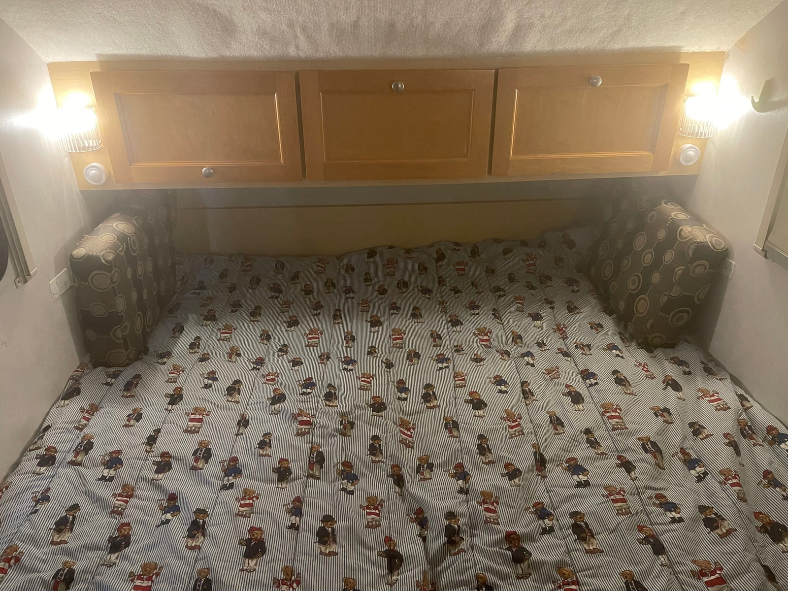 Inside an RV rental: a cozy bed with teddy bear striped comforter, overhead cabinets, and reading lights.