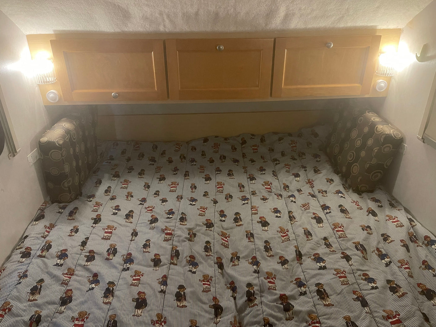 Inside an RV rental: a cozy bed with teddy bear striped comforter, overhead cabinets, and reading lights.