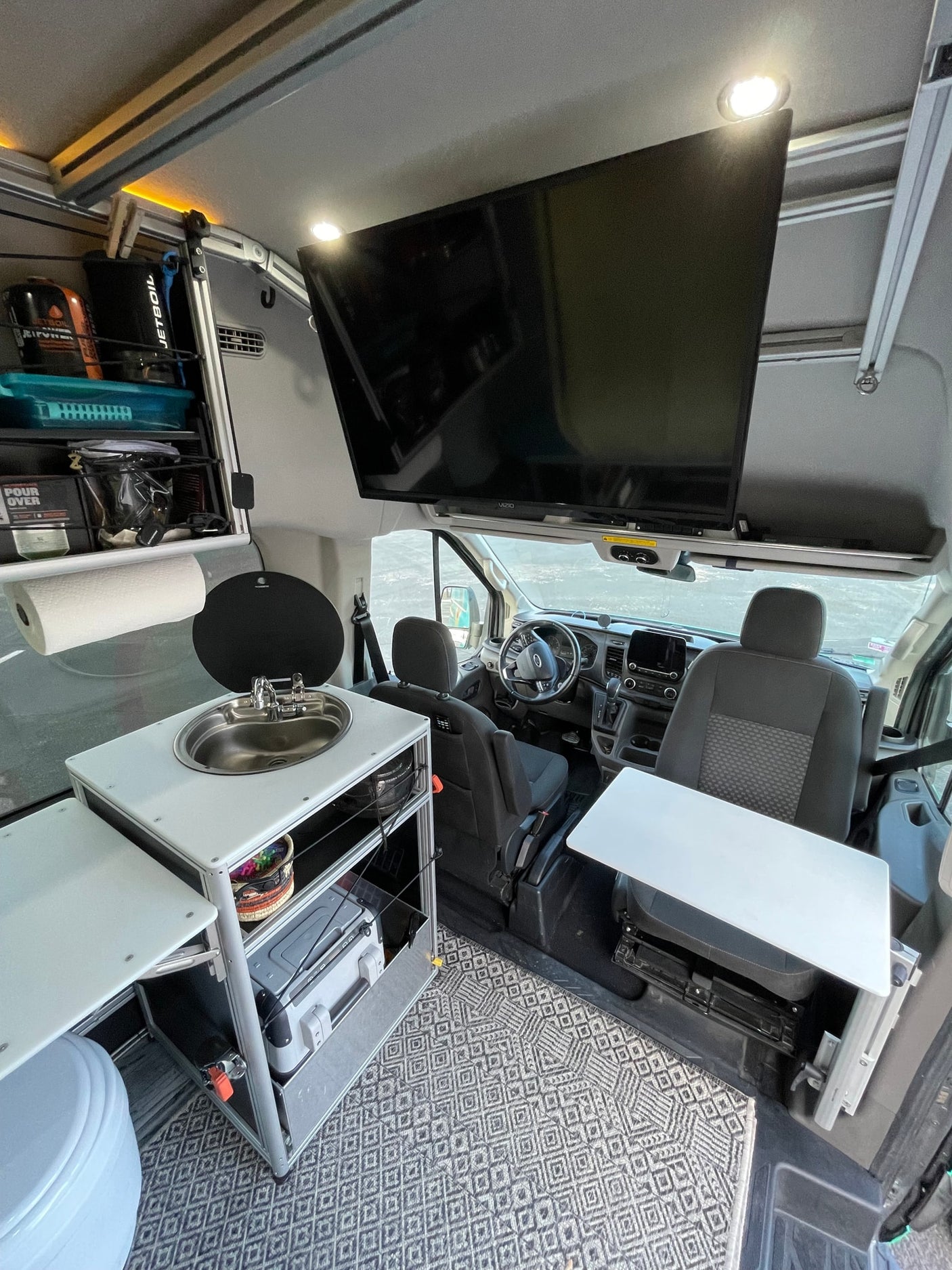 Inside a modern RV rental, featuring a compact kitchenette, large TV, swivel table, and the driver's area.