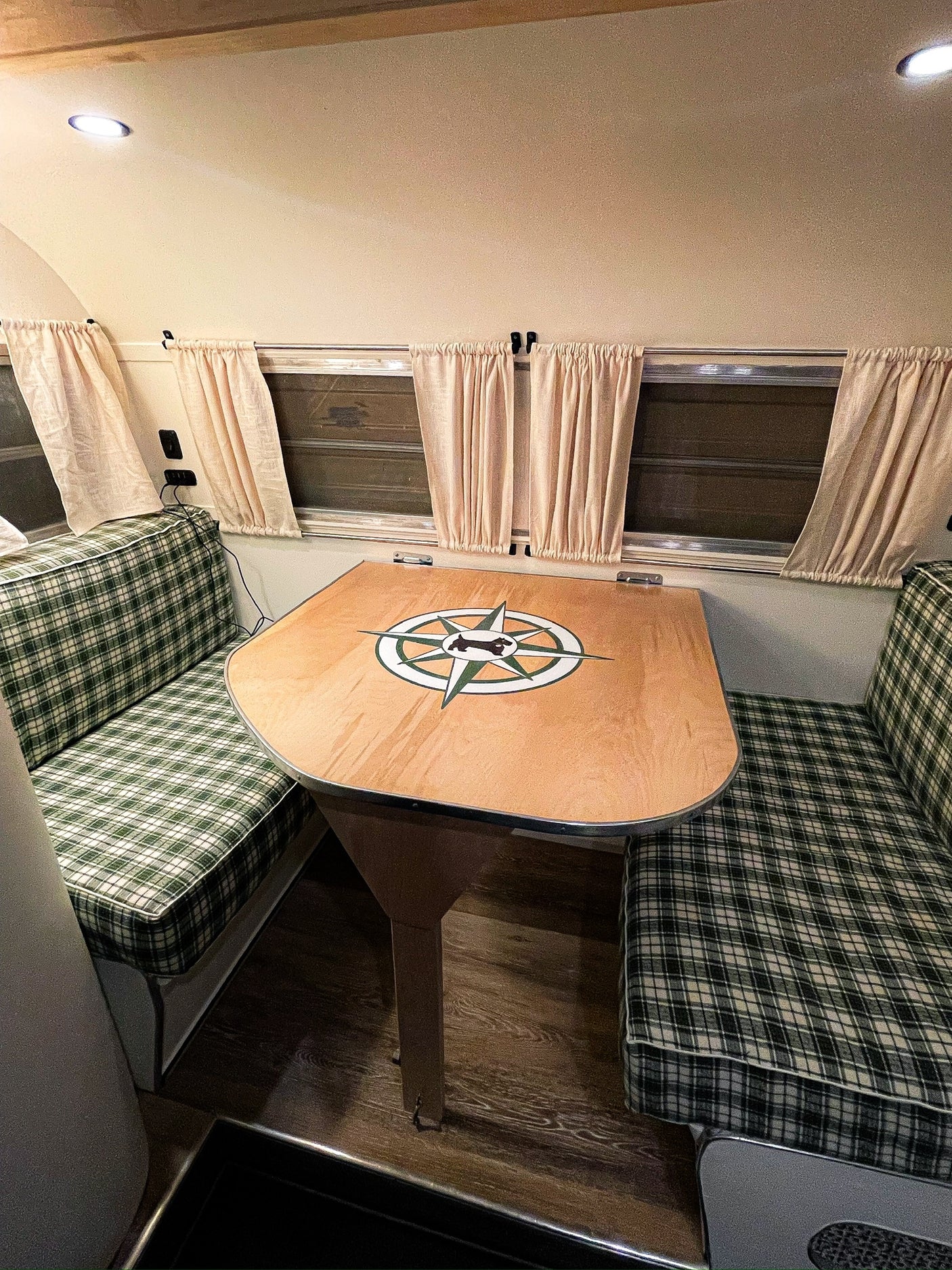 Inside a cozy RV rental: green plaid dinette seating, wooden table with compass design, and windows with curtains.