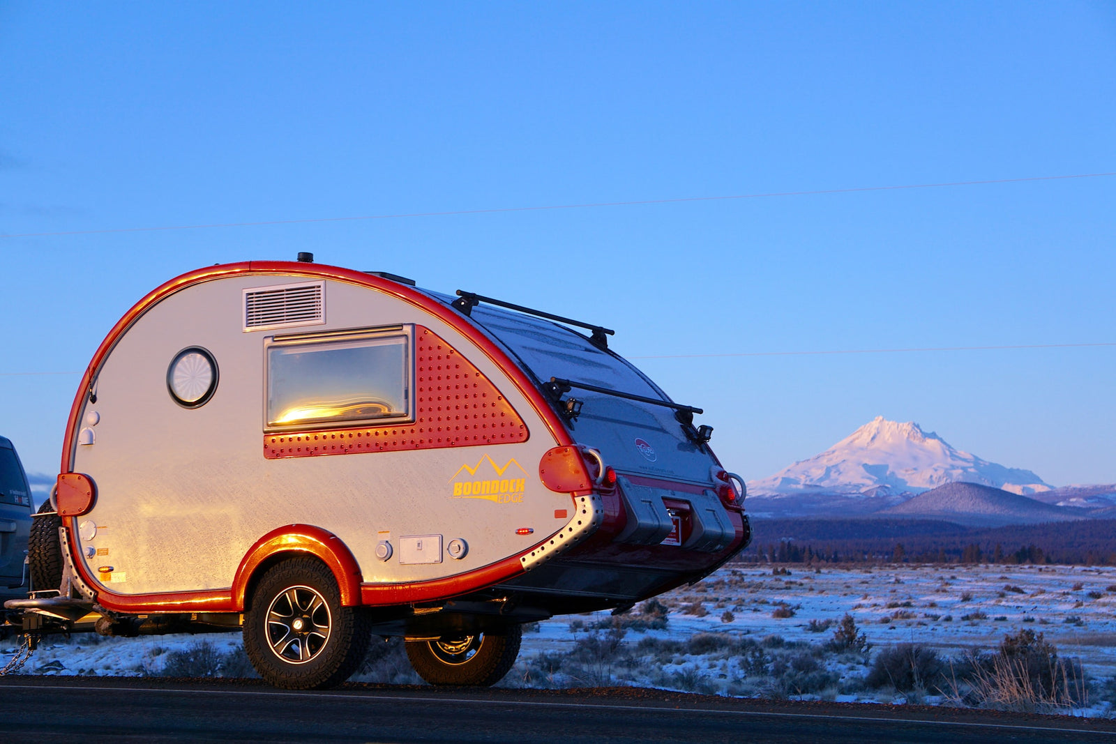 Stylish teardrop RV rental parked roadside in snowy landscape with a majestic snow-capped mountain backdrop.