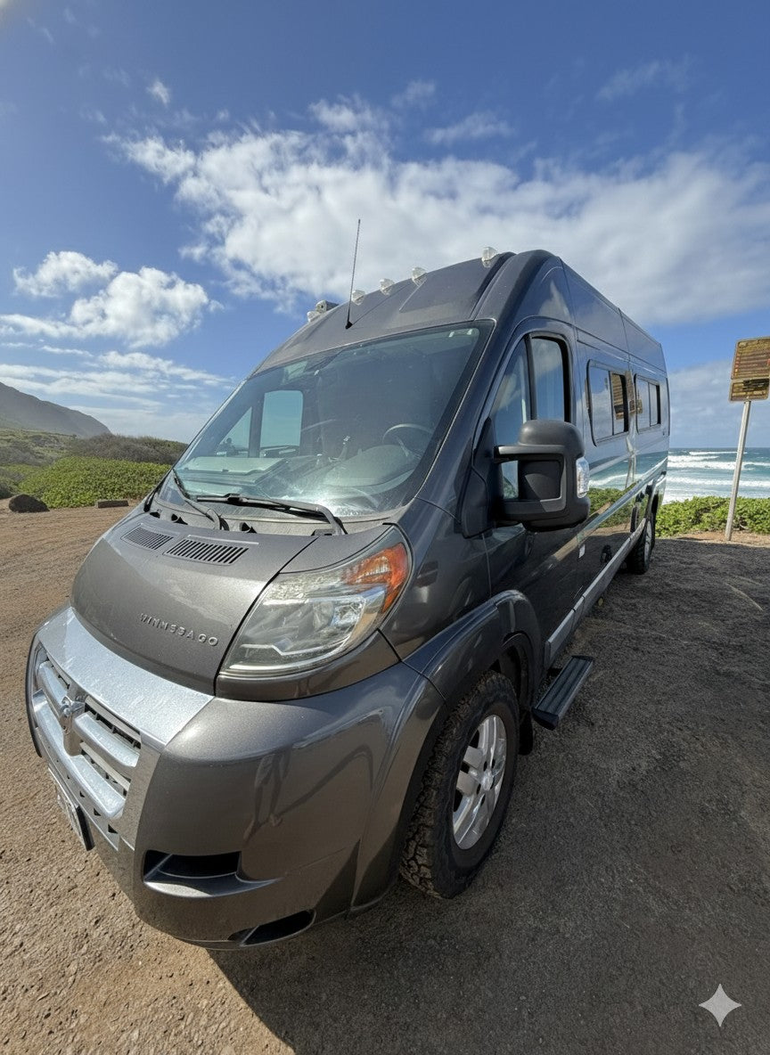 A gray Winnebago campervan (rv rental) parked on a dirt road next to a sunny beach with ocean waves under a blue sky.