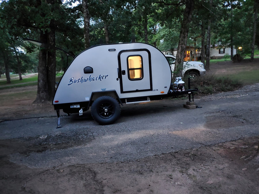 A Bushwhacker teardrop RV rental, a compact travel trailer with an illuminated interior, parked on a gravel path at dusk.