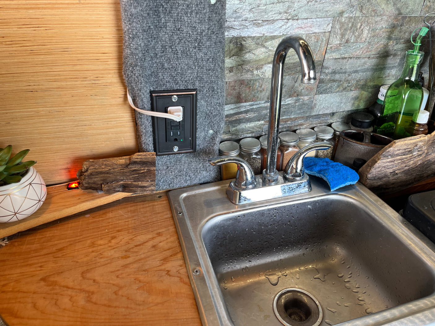 RV rental's compact kitchen setup: stainless sink, chrome faucet, spice jars, and a power outlet on carpeted wall.