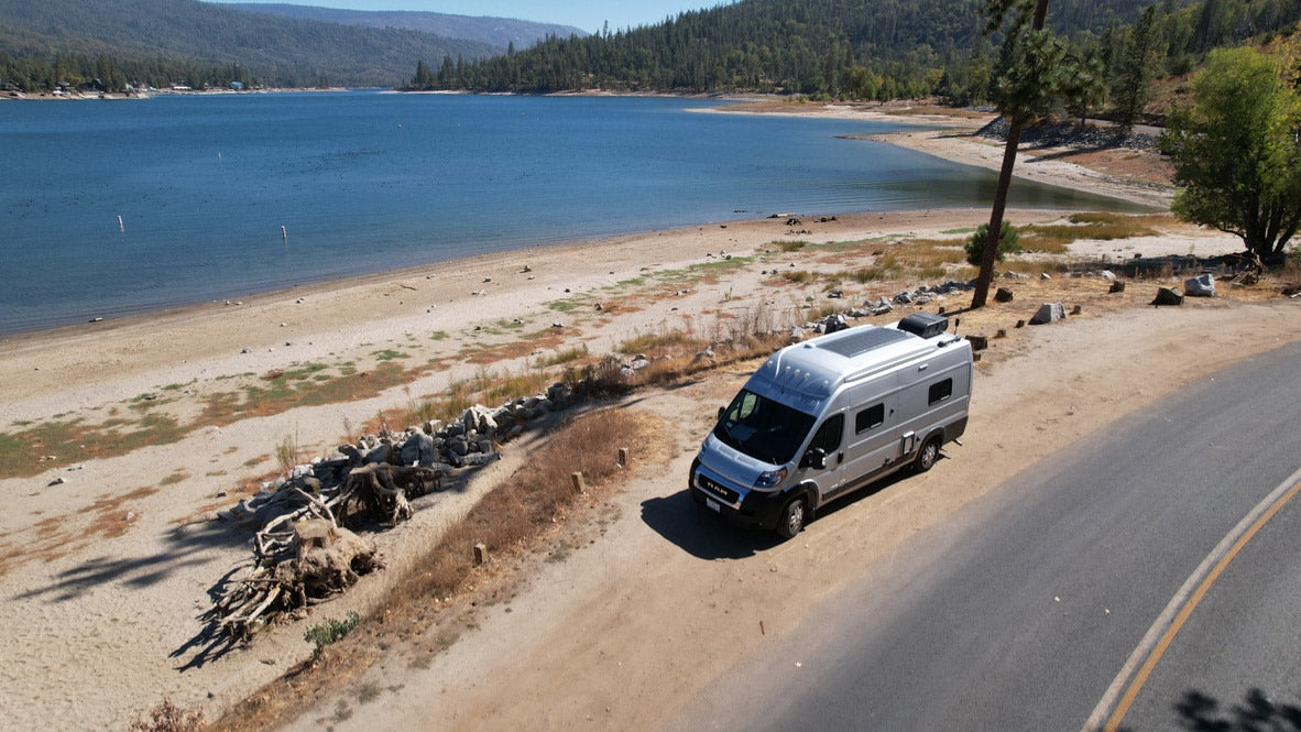 An RV rental offers prime views. A silver camper van is parked on a dirt pull-off overlooking a large lake and sandy beach amidst mountains.