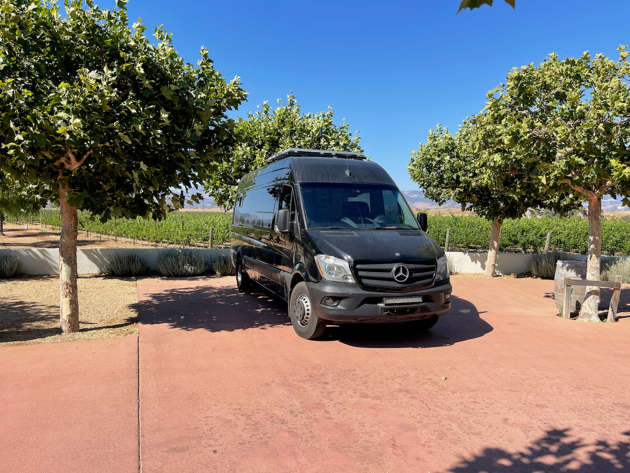 A black Mercedes Sprinter rv rental is parked on a red patio at a sunny vineyard, surrounded by trees and distant hills.