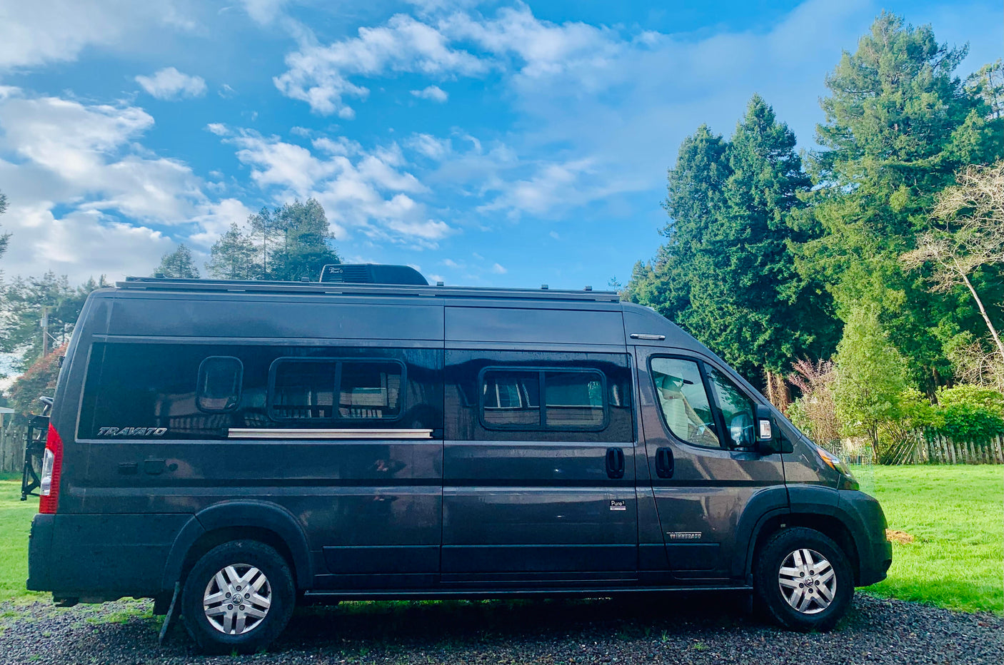 Dark gray Winnebago Travato RV rental, a modern camper van, parked on gravel next to green trees under a blue sky.