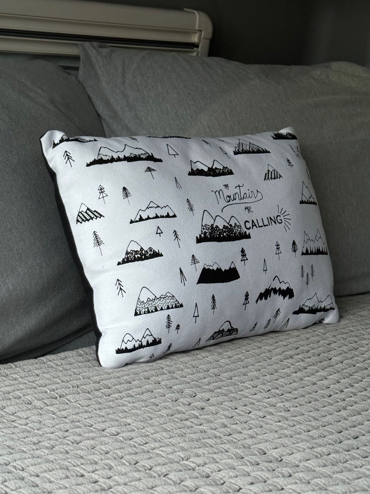 Cozy RV rental bed with a mountain-themed pillow, "The Mountains Are CALLING," and soft grey bedding.