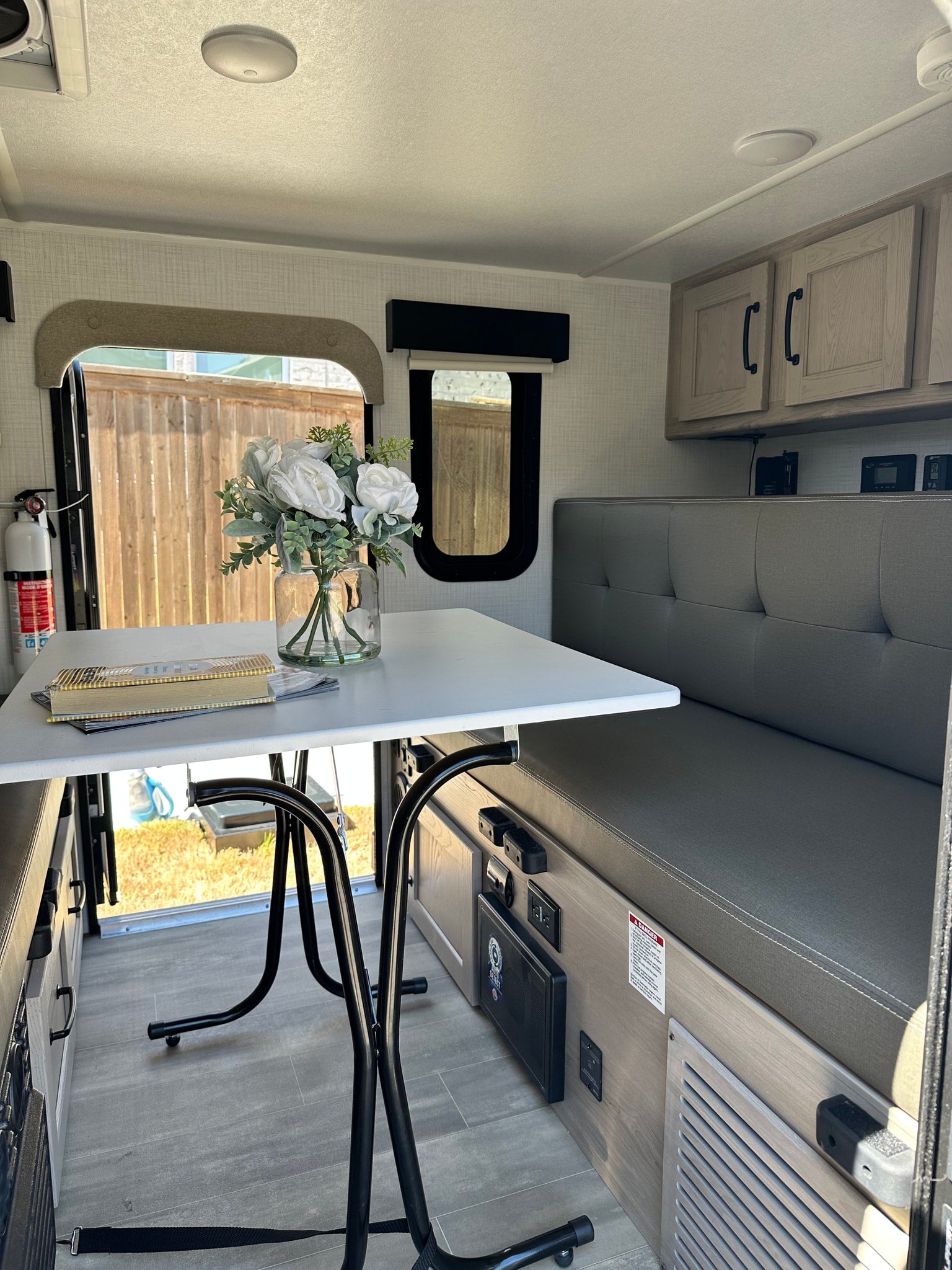 Modern rv rental dinette area with white table, gray bench, and overhead cabinets. Window shows outdoor fence.