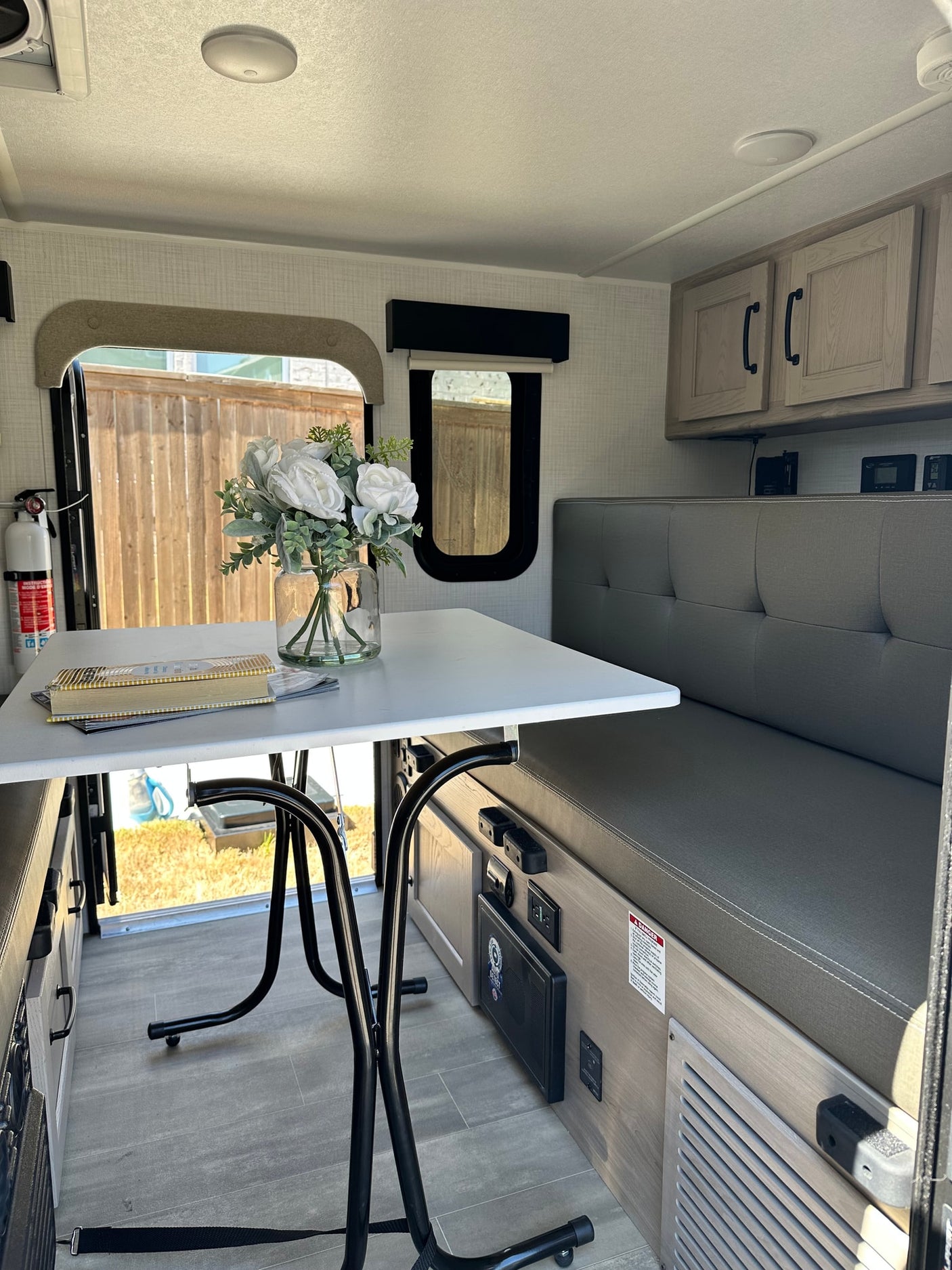 Modern rv rental dinette area with white table, gray bench, and overhead cabinets. Window shows outdoor fence.