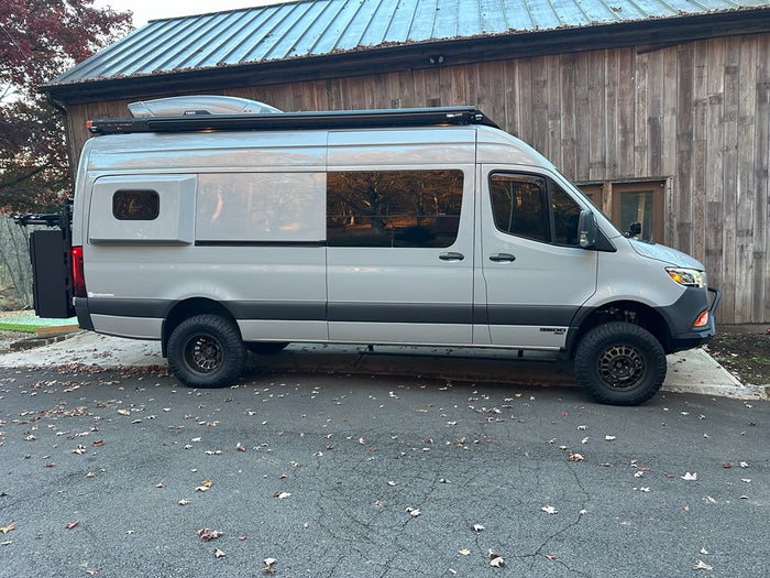 parked on a gravel path in a forest.|This light grey Sprinter camper van