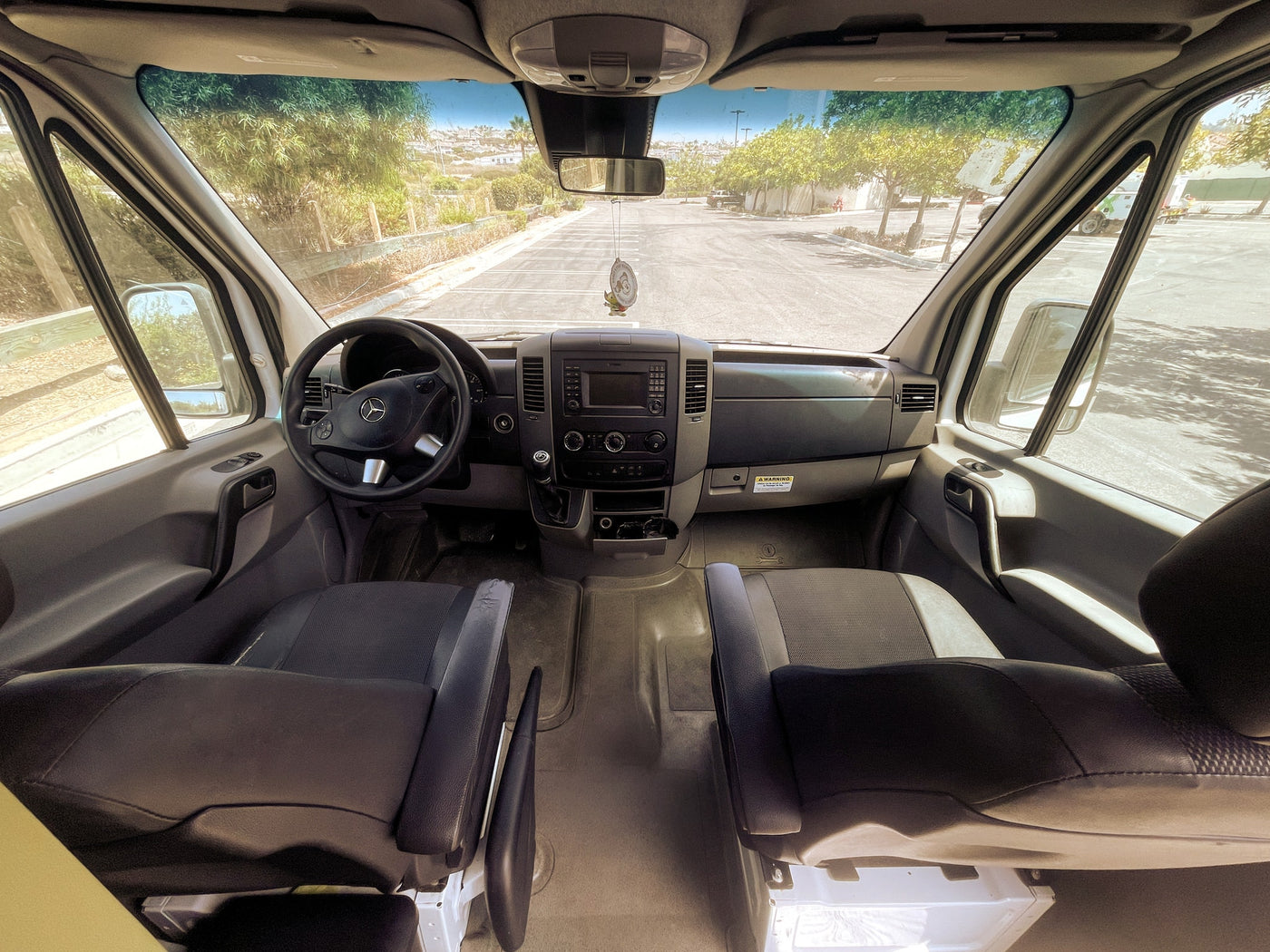 Get a feel for your RV rental! View from inside the van showing the cockpit, driver/passenger seats, and the sunny parking lot ahead. — photo 14