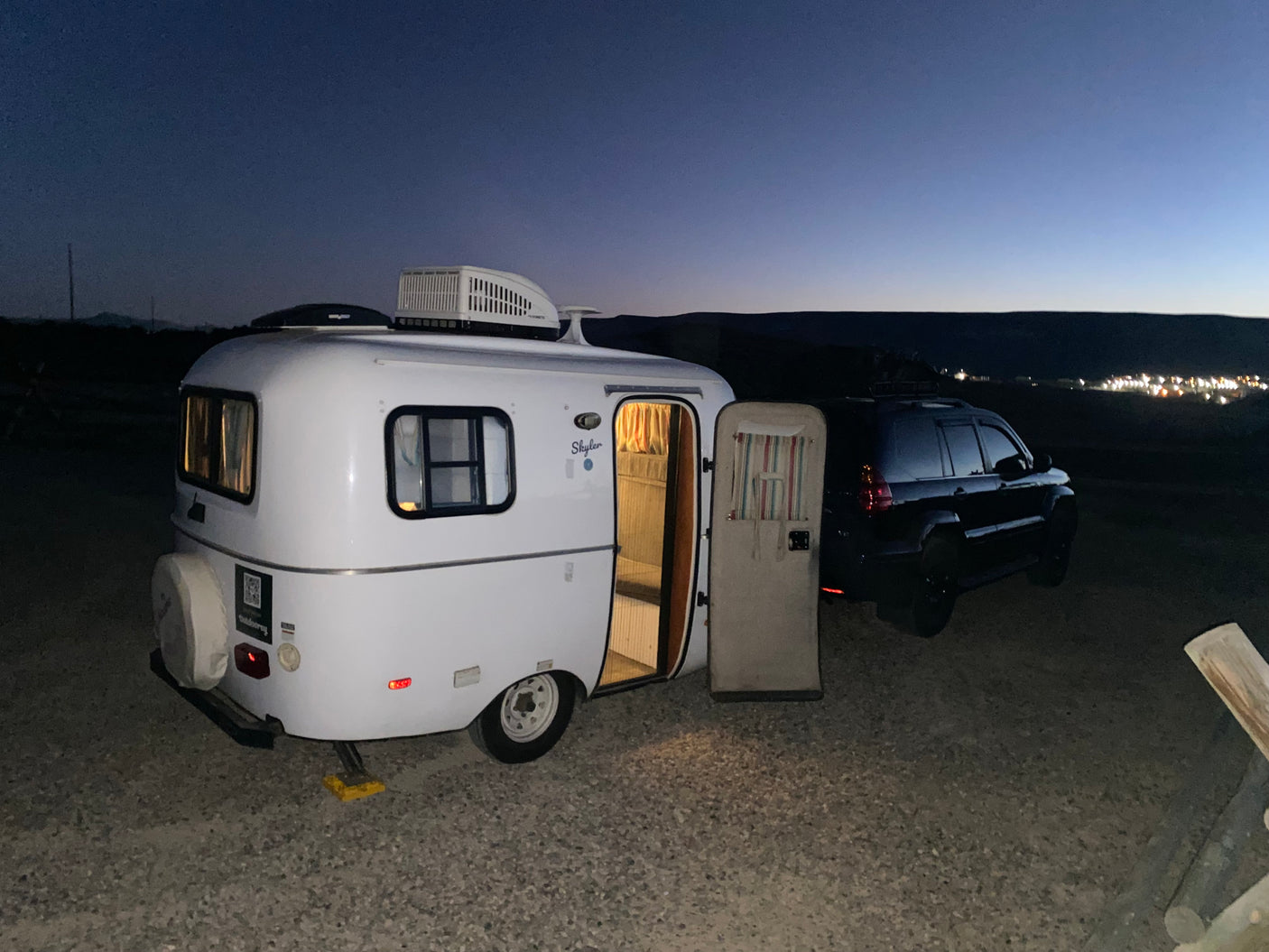 A white Skyler teardrop RV rental with its door open, hitched to a dark SUV, parked on gravel at dusk with distant city lights.