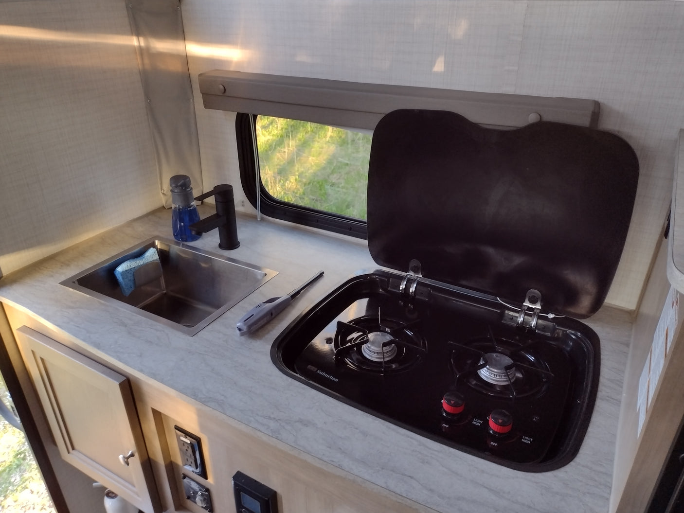 Compact RV rental kitchen with sink, faucet, 2-burner stove (cover open), and a window view of green grass. Ready for meals on the go!