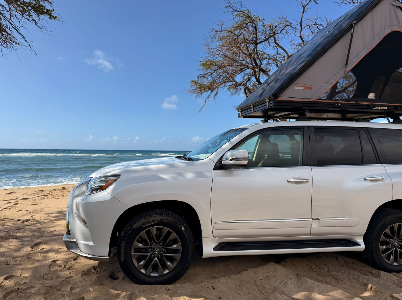 Adventure-ready RV rental: White SUV with a Roofnest rooftop tent on a sandy beach next to the ocean under a clear blue sky.