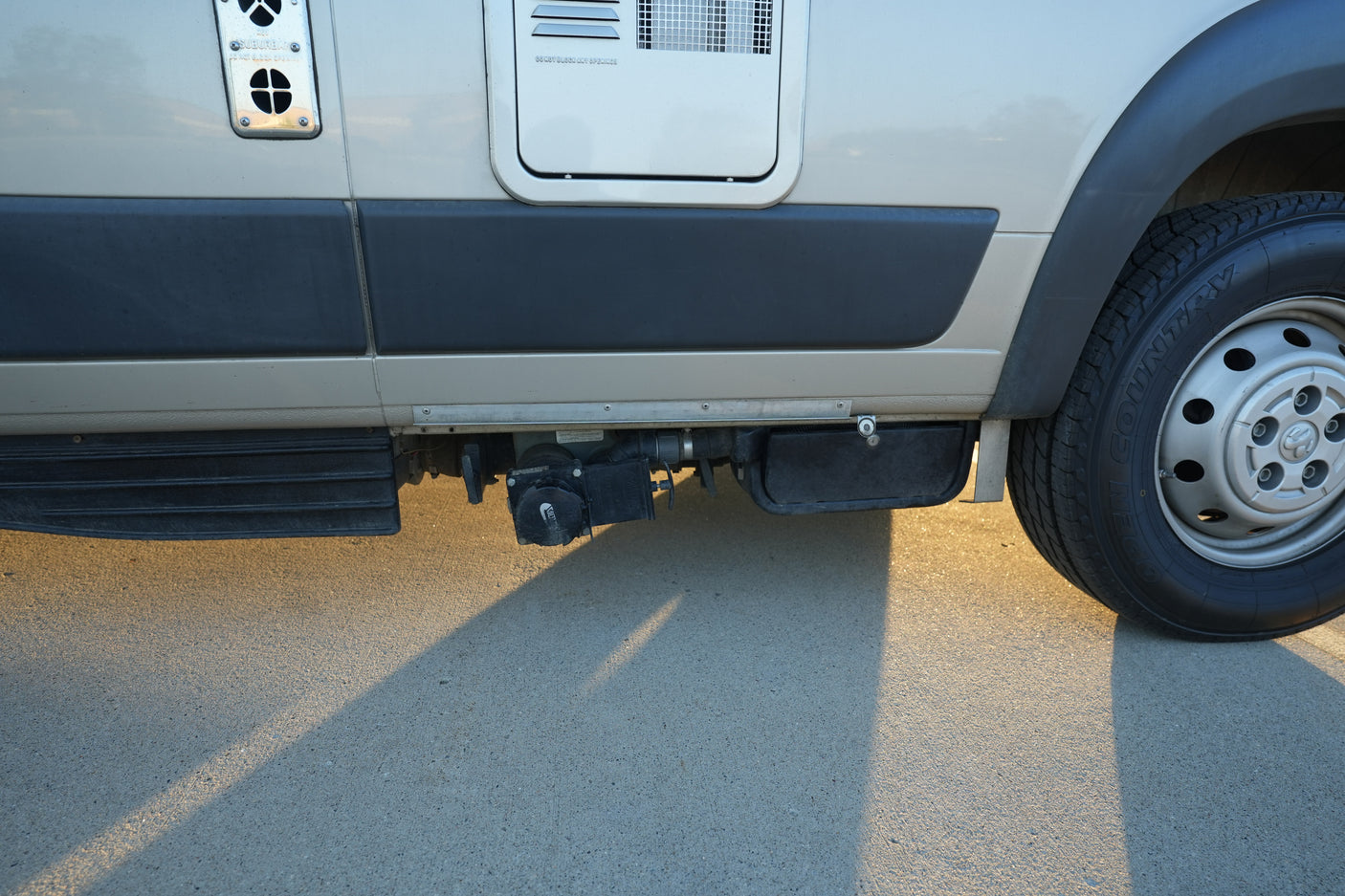 RV rental: Side view showing waste tank plumbing, tire, and appliance vents. Sunlit paved surface below.