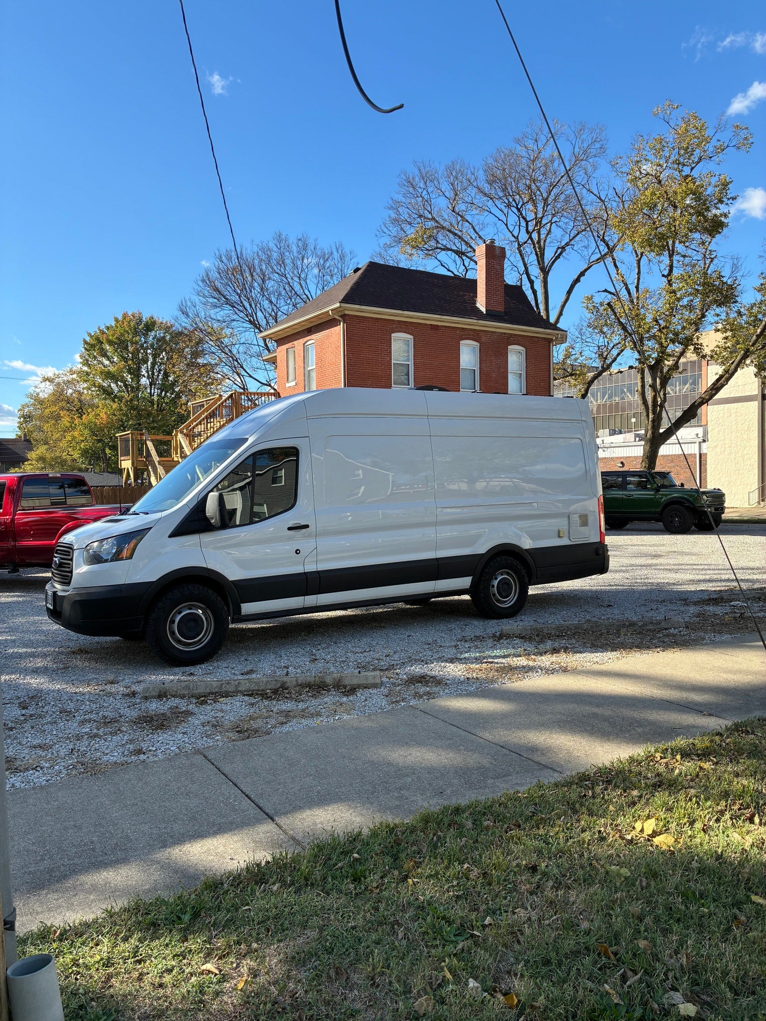 White Ford Transit van, a popular RV rental option, parked on gravel. A red truck, green Bronco, brick house, and trees are in view.