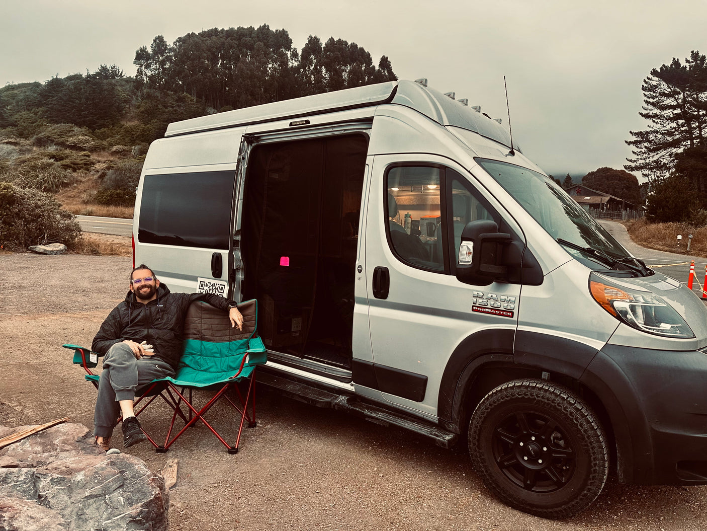 Man relaxes in a chair beside a silver Ram ProMaster RV rental on a gravel road, surrounded by trees.
