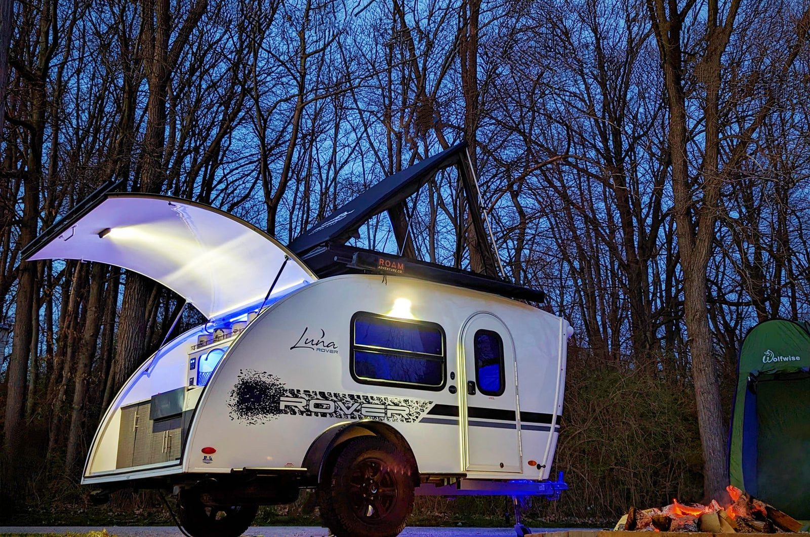 Enjoy the outdoors! This Luna Rover teardrop RV rental, fully lit, awaits adventure with a cozy campfire and tent at dusk.