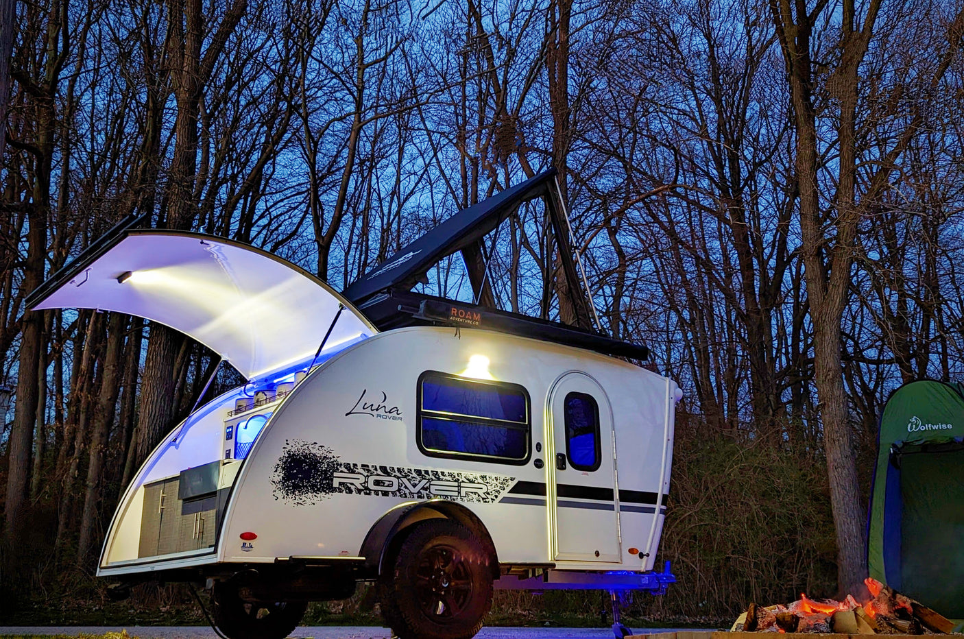 Enjoy the outdoors! This Luna Rover teardrop RV rental, fully lit, awaits adventure with a cozy campfire and tent at dusk.