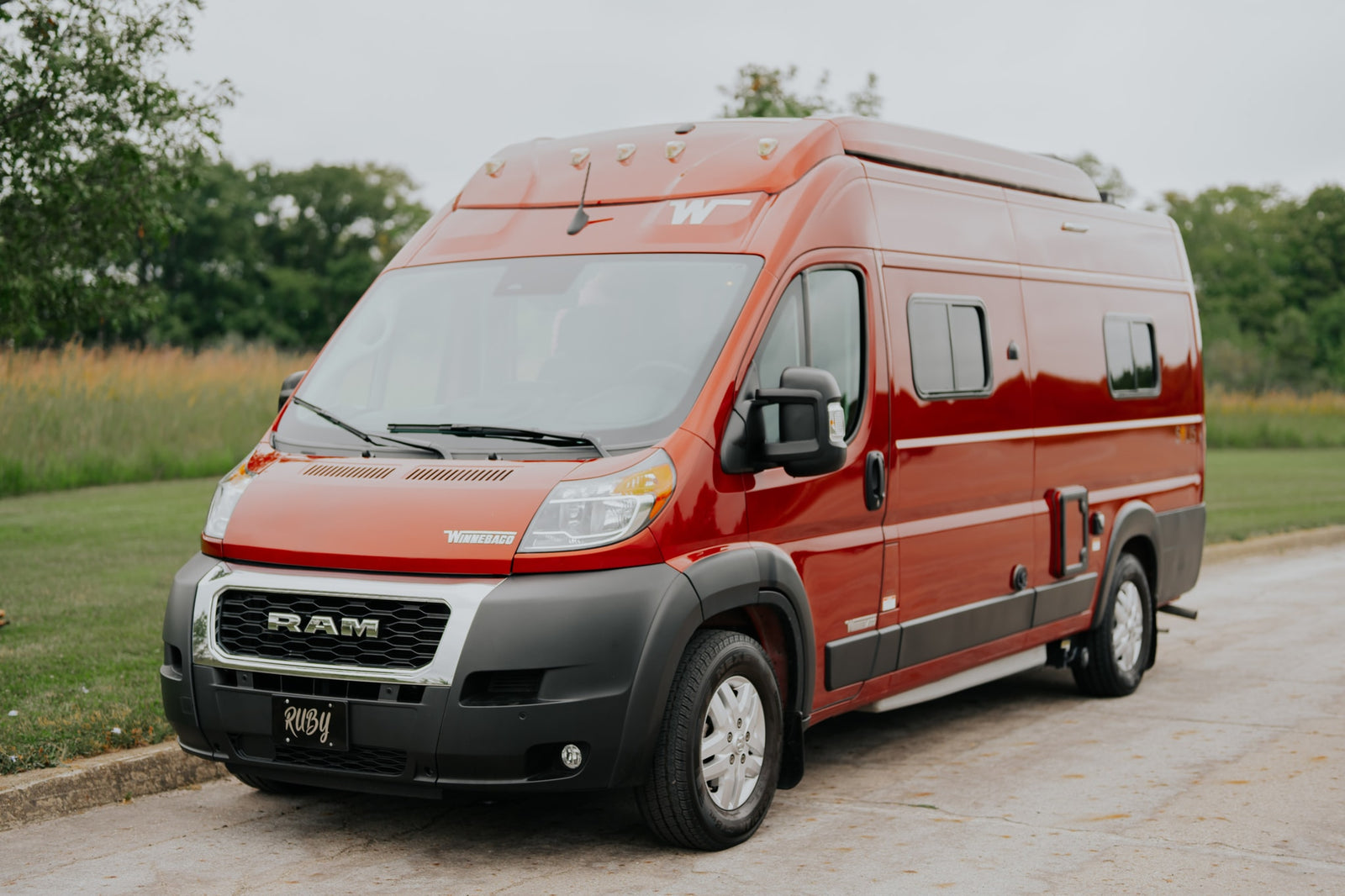 A vibrant red Winnebago camper van, available for rv rental, parked on asphalt with green foliage in the background.