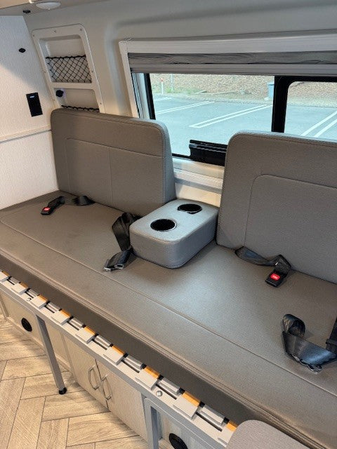 This RV rental interior features a gray bench seat with seatbelts and cup holders. Under-seat storage and a window are visible.