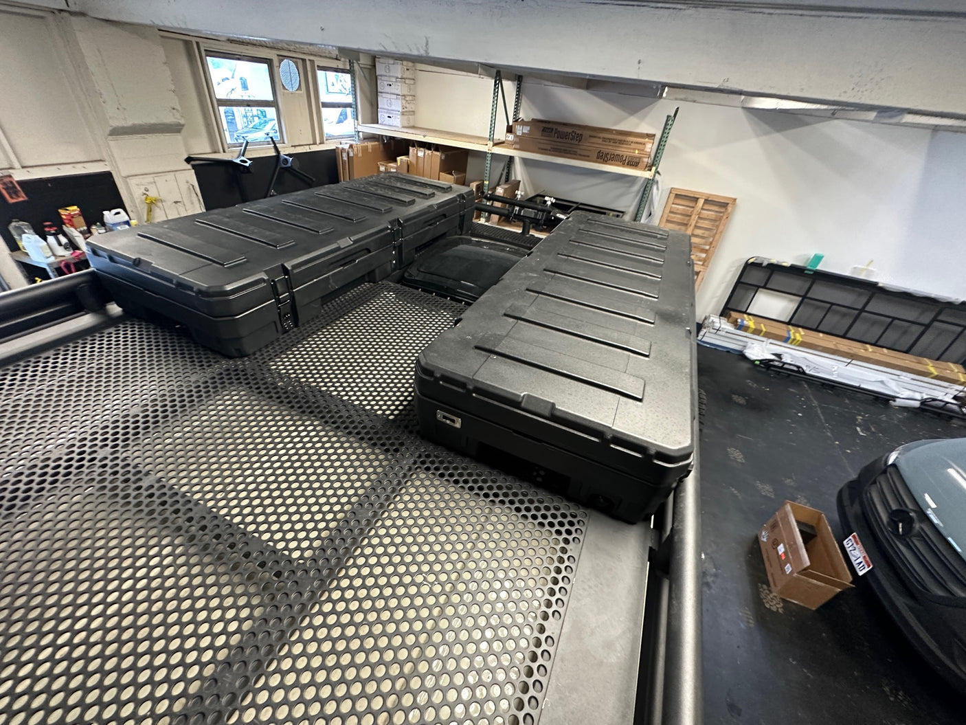 Large black storage cases are mounted on a perforated roof rack, possibly for an RV rental or adventure vehicle, in a workshop setting.