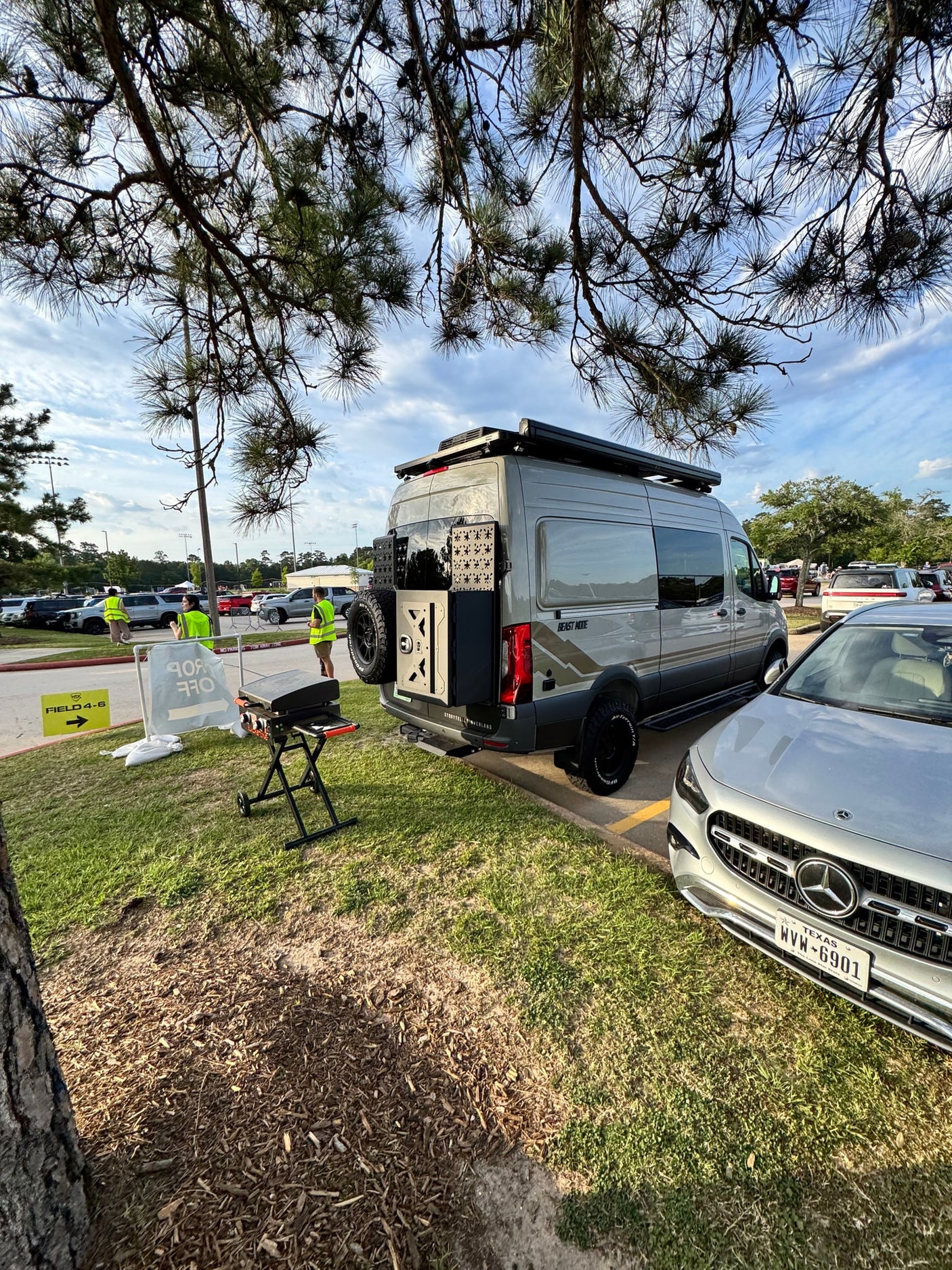 A grey Storyteller Overland 'Beast Mode' campervan next to a Mercedes-Benz car. RV rental drop-off area with a portable grill and event signs.