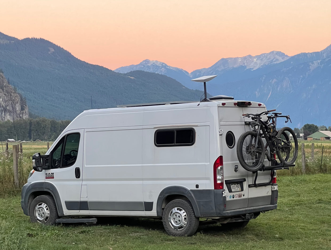 RV rental with bikes and Starlink dish in a grassy field, backed by snow-capped mountains under an orange-pink sunset sky.