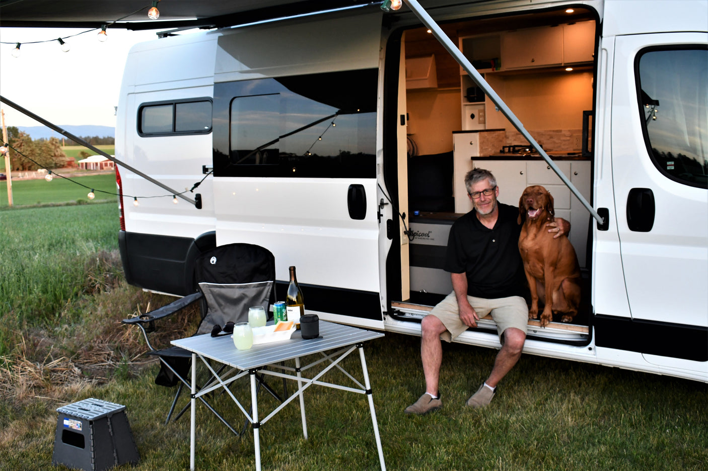 RV rental scene: A man and his dog relax at sunset by their camper van with an awning, outdoor table, and string lights.