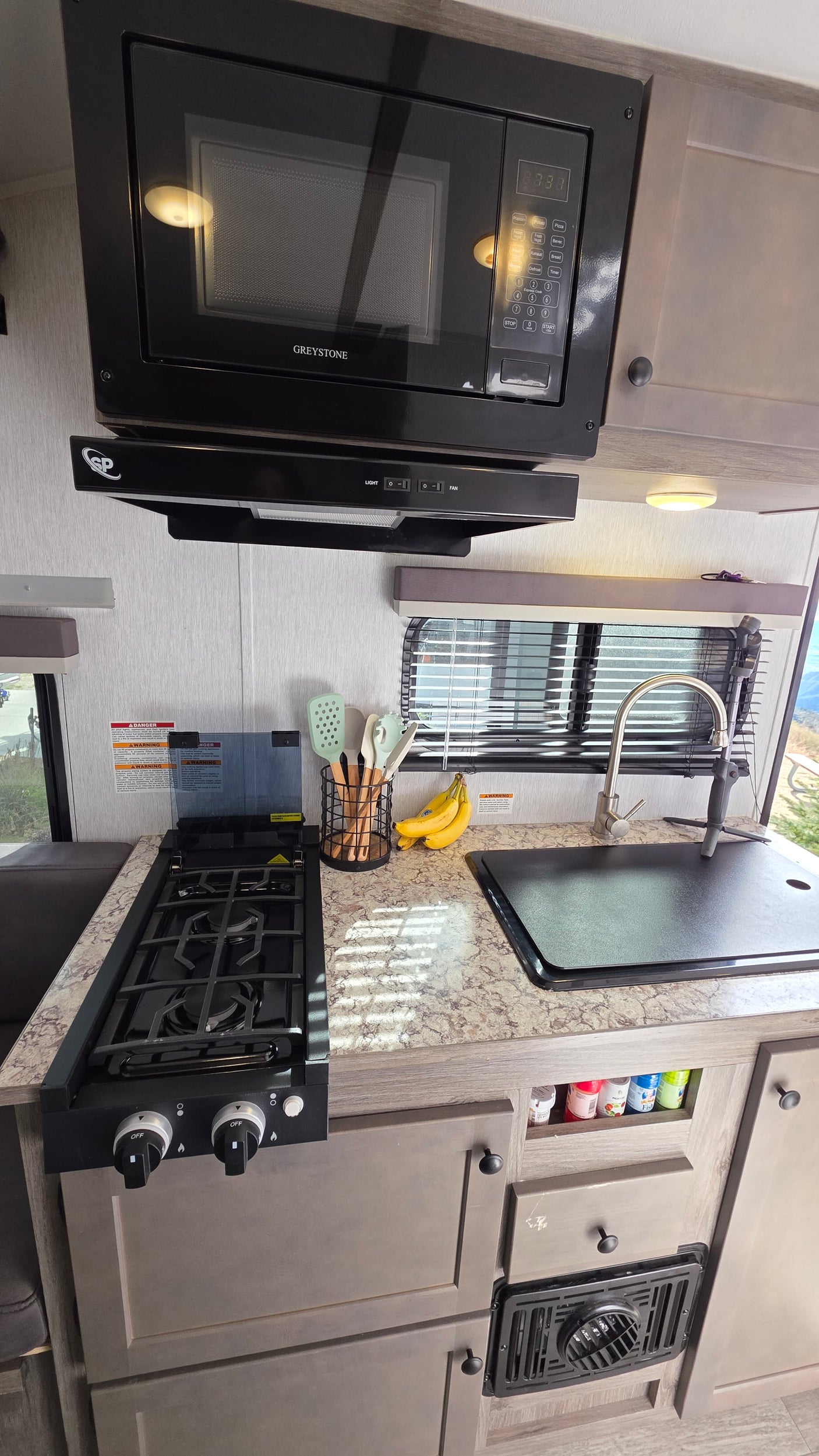 An RV rental kitchen, equipped with a Greystone microwave, range hood, gas stove, covered sink, and counter space.