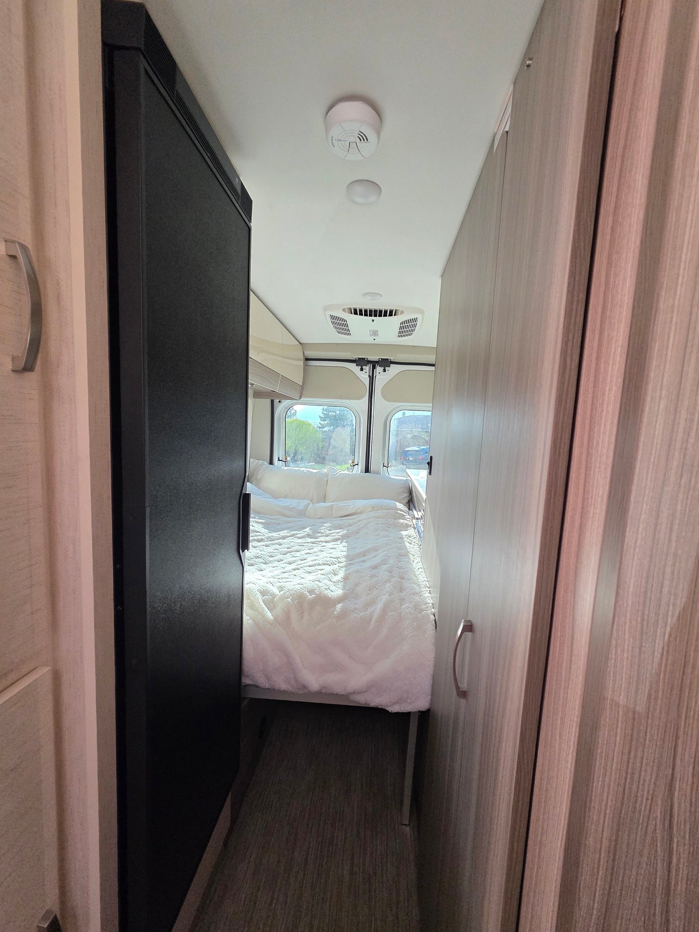 View down a narrow RV rental interior showing a cozy white bed at the end, flanked by a refrigerator and wood-grain cabinets.