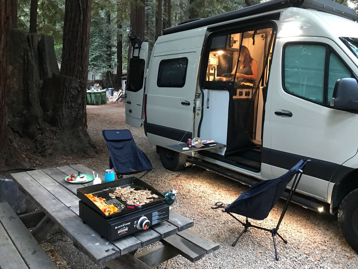 RV rental camp life: A person cooks dinner on a griddle outside their camper van amidst tall forest trees.|Bright sun illuminates an RV rental Sprinter van on a gravel campsite amidst tall forest trees