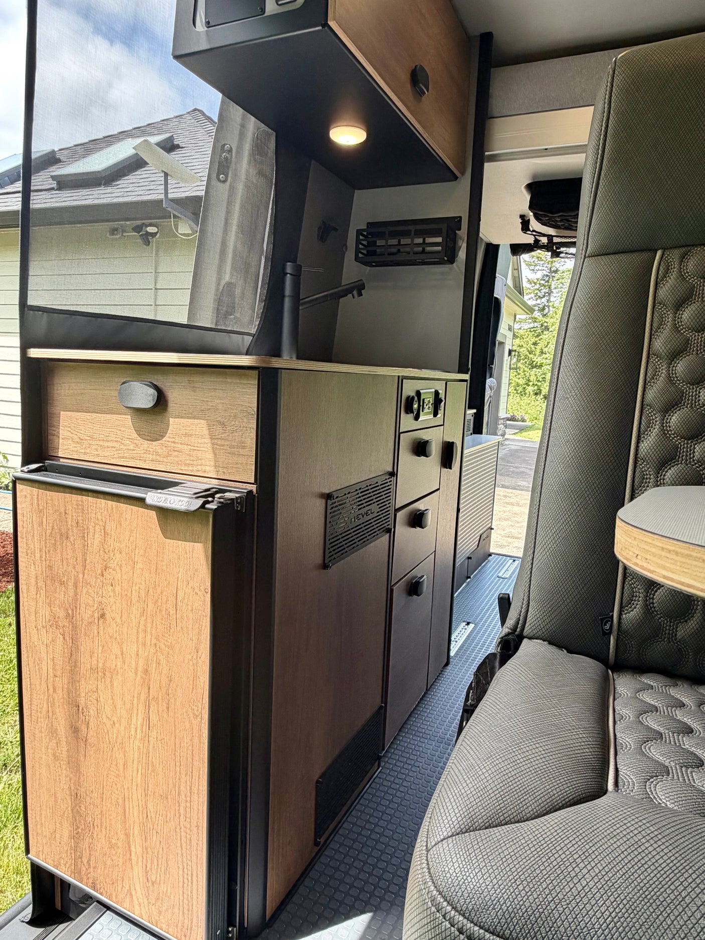 Modern rv rental interior with kitchen, fridge, cabinets, and seating. View outside through screen shows house and sky.