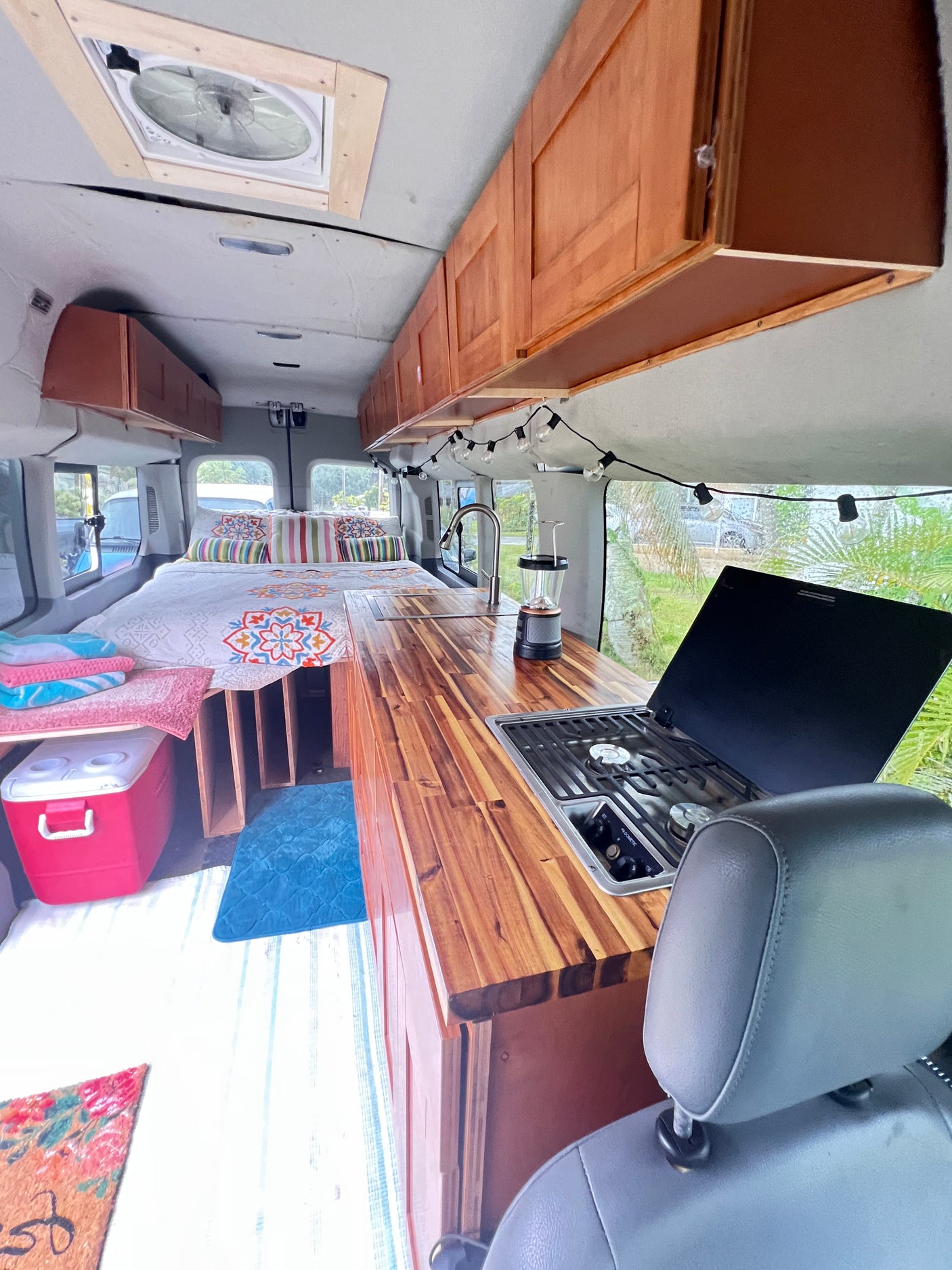 RV rental interior: cozy bed, kitchen with sink & stove, wood counters, and overhead storage.
