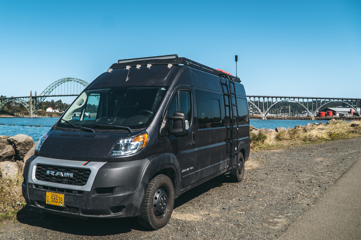 Black Ram ProMaster camper van, likely an RV rental, parked on a gravel road by a scenic waterfront with a large bridge.