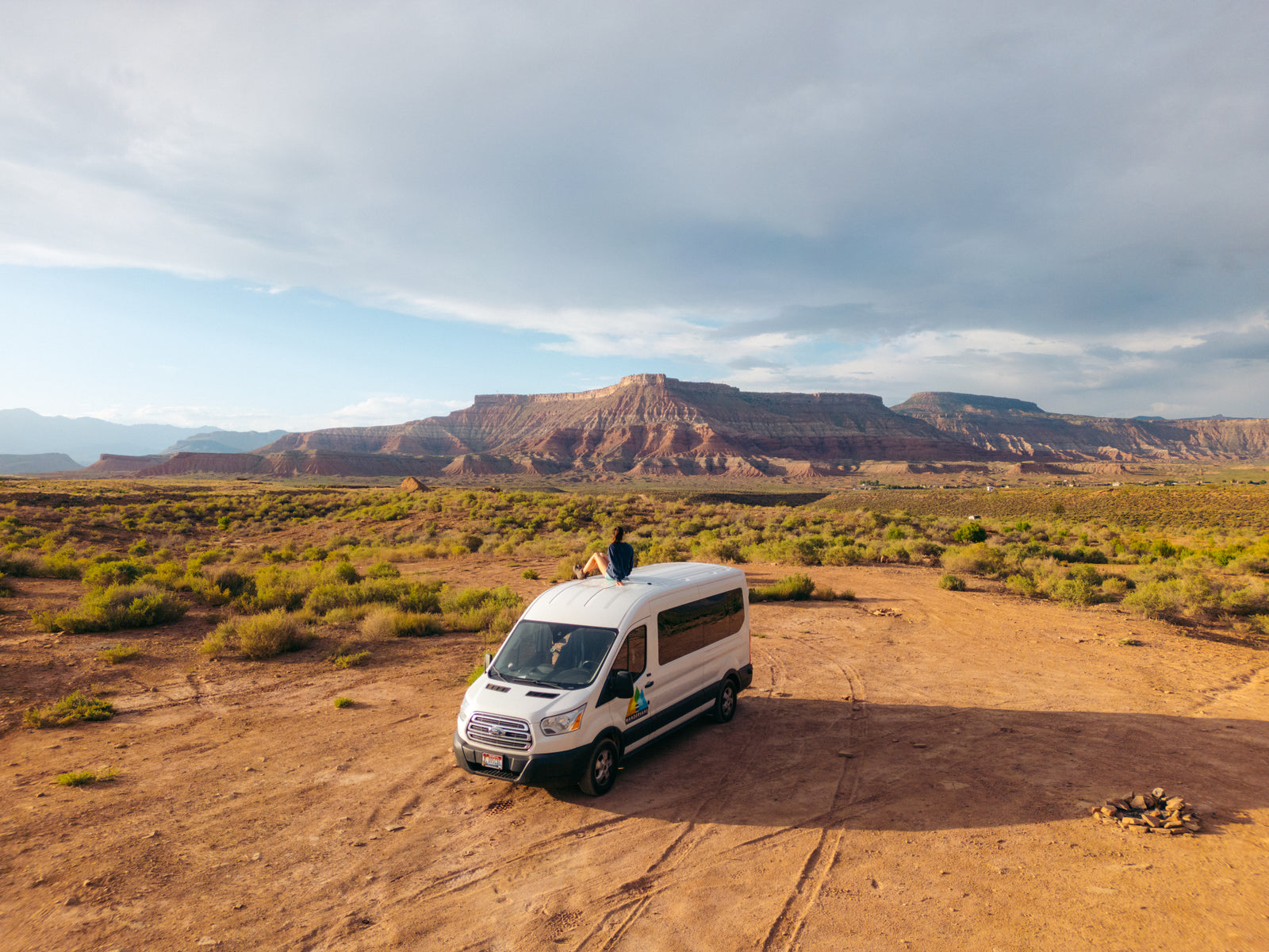 Scenic rv rental: A person sits atop a camper van, overlooking a vast desert landscape with red rock mountains under a cloudy sky.