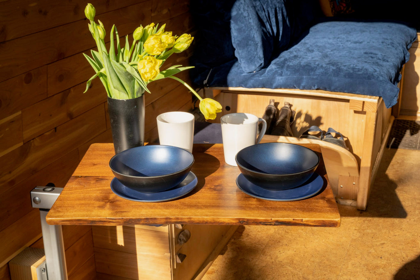 Sunlit RV rental interior: wooden walls, blue bedding, and a cozy table set with blue dishes, white mugs, and yellow tulips.