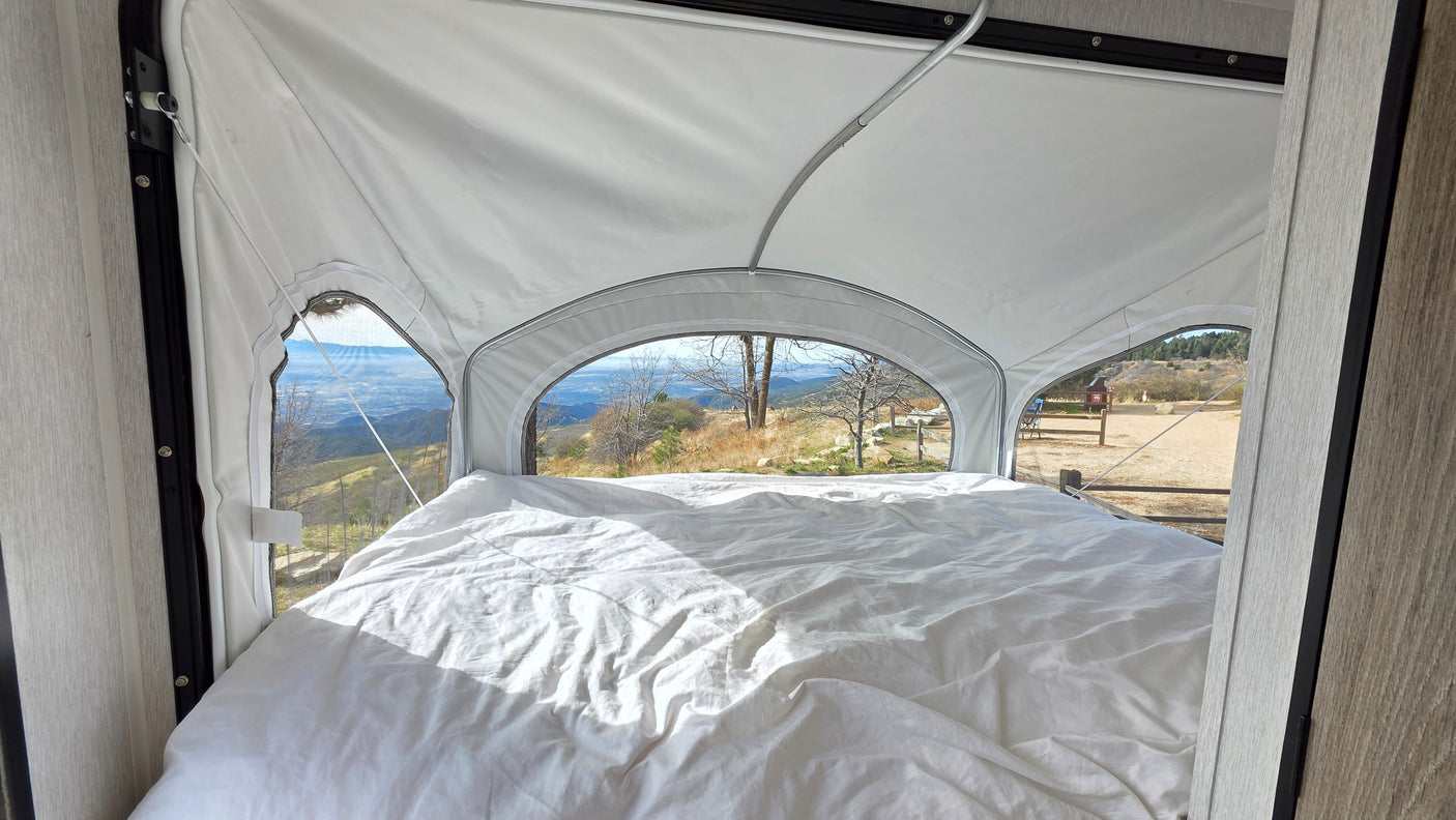 Inside an RV rental, a comfy bed faces panoramic mountain views from a pop-up tent extension. A campsite is visible outside.
