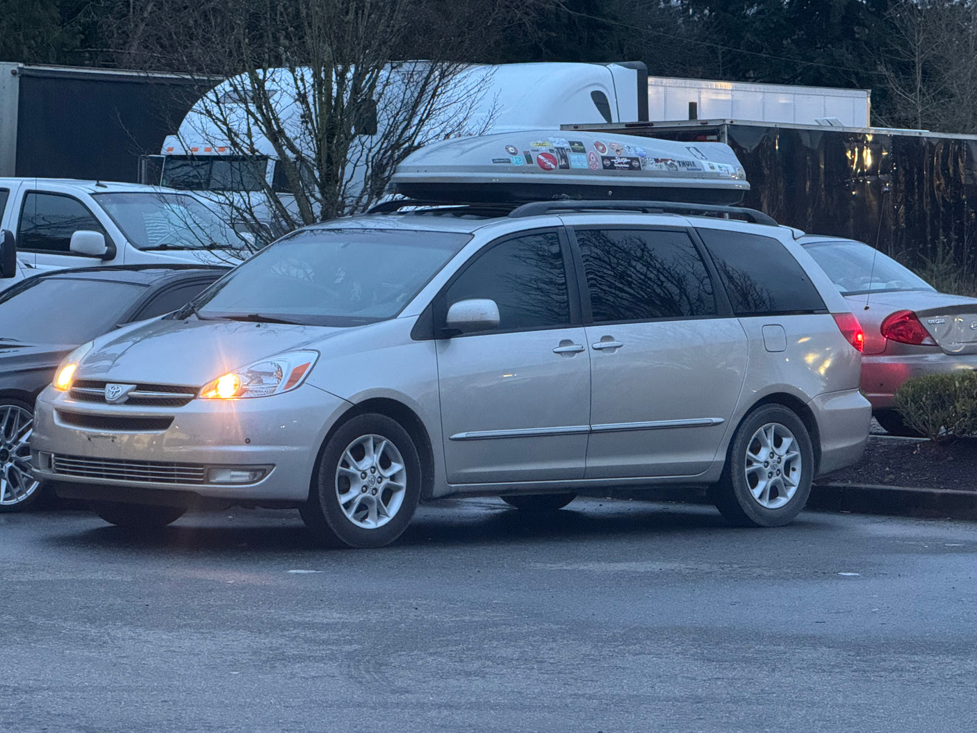 Silver minivan with a roof cargo box and bright headlights, parked near commercial trailers, suggesting an RV rental area.