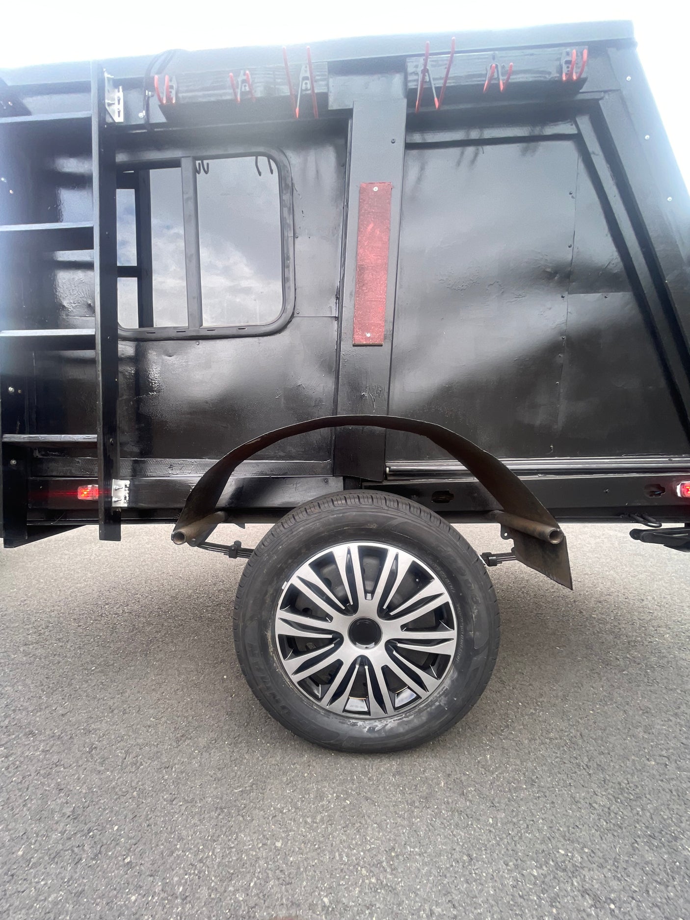 Side view of a black RV rental trailer, featuring a ladder, window, two-tone spoked wheel, and red clips for equipment.