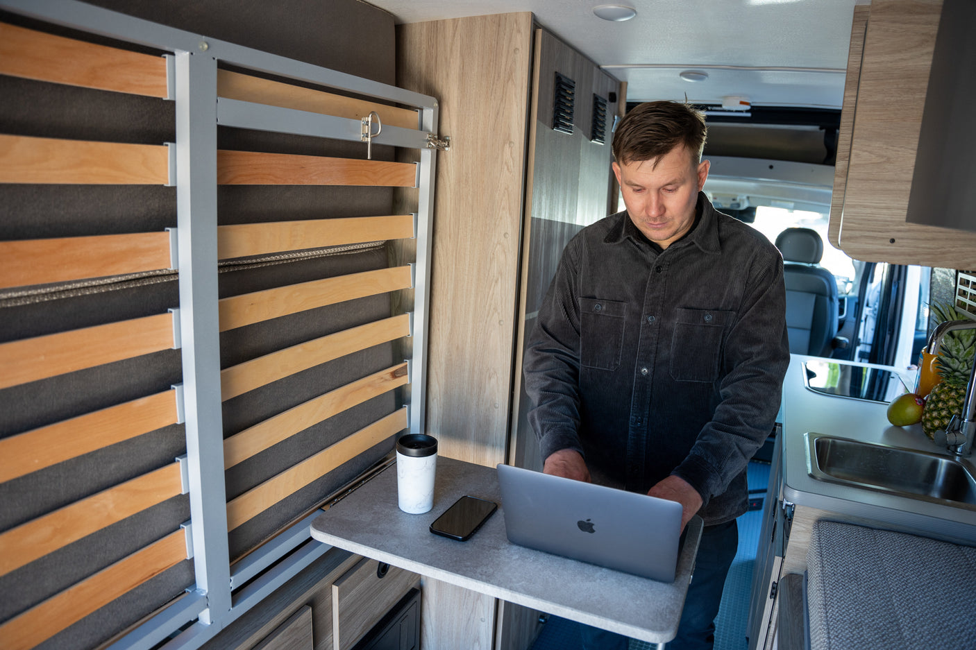 A man works on a laptop in a modern RV rental, featuring a compact kitchen, space-saving bed, and mobile office setup.